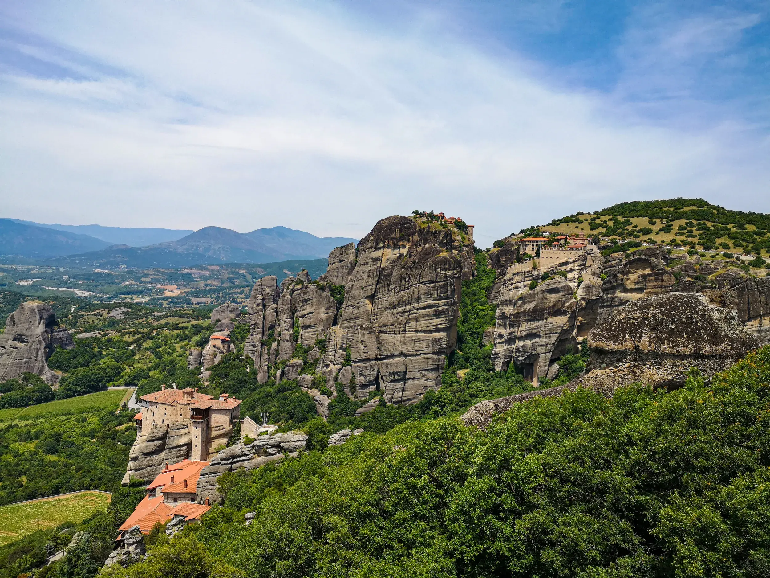 Blick von einer Wanderung auf die Meteora Klöster, die auf steilen Felsen über einem grünen Tal in Griechenland thronen.