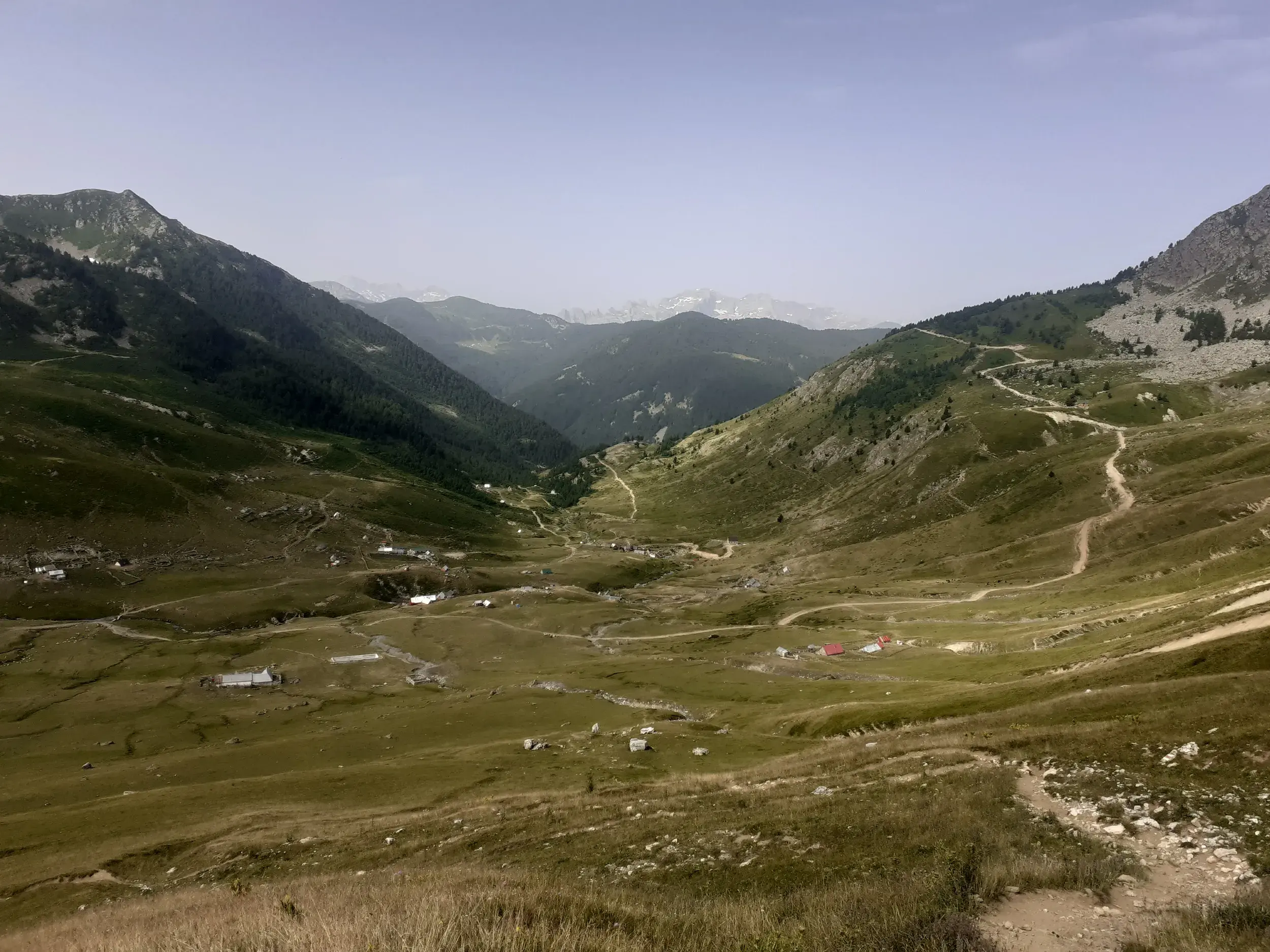Blick über ein weites, grünes Hochtal mit Berghütten und einem schmalen Wanderweg auf dem Peaks of the Balkans Trail.