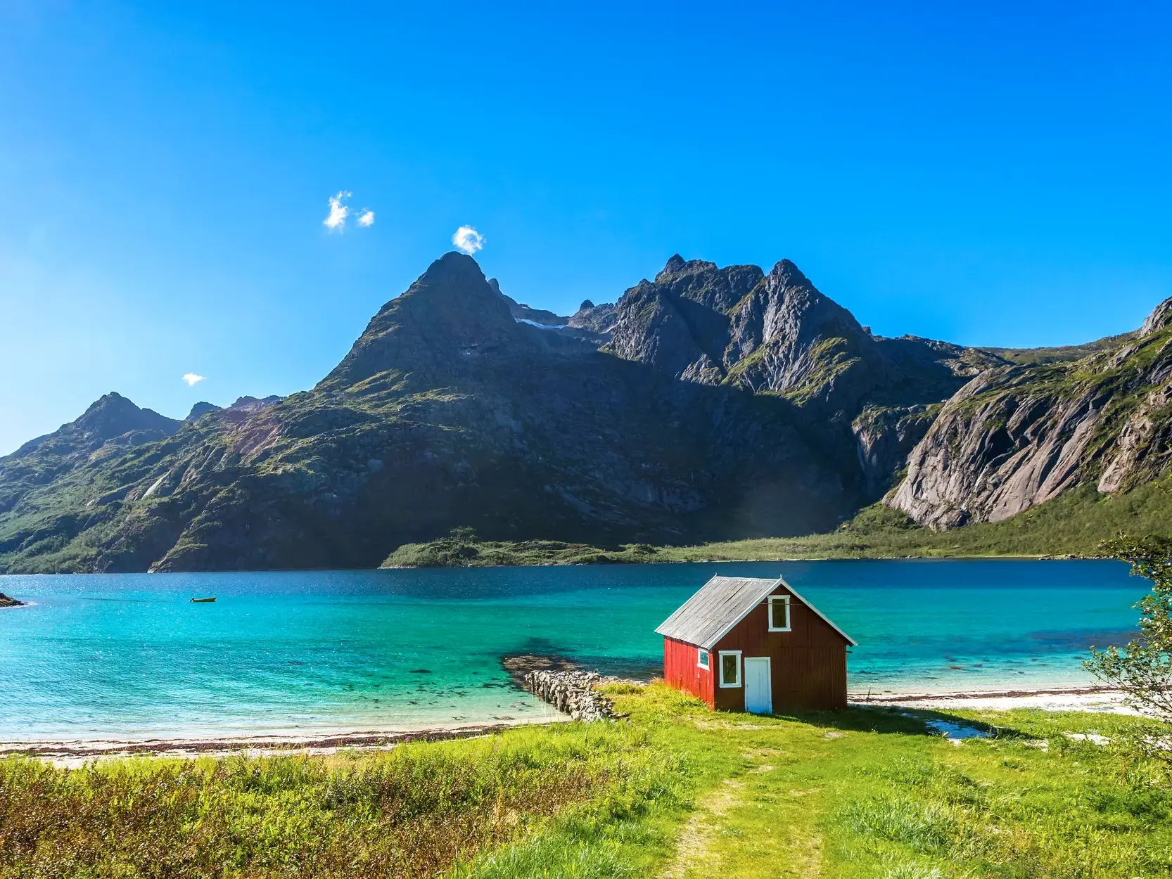 Idyllische rote Fischerhütte am Ufer eines Fjords auf den Lofoten mit türkisem Wasser und imposanter Bergkulisse.