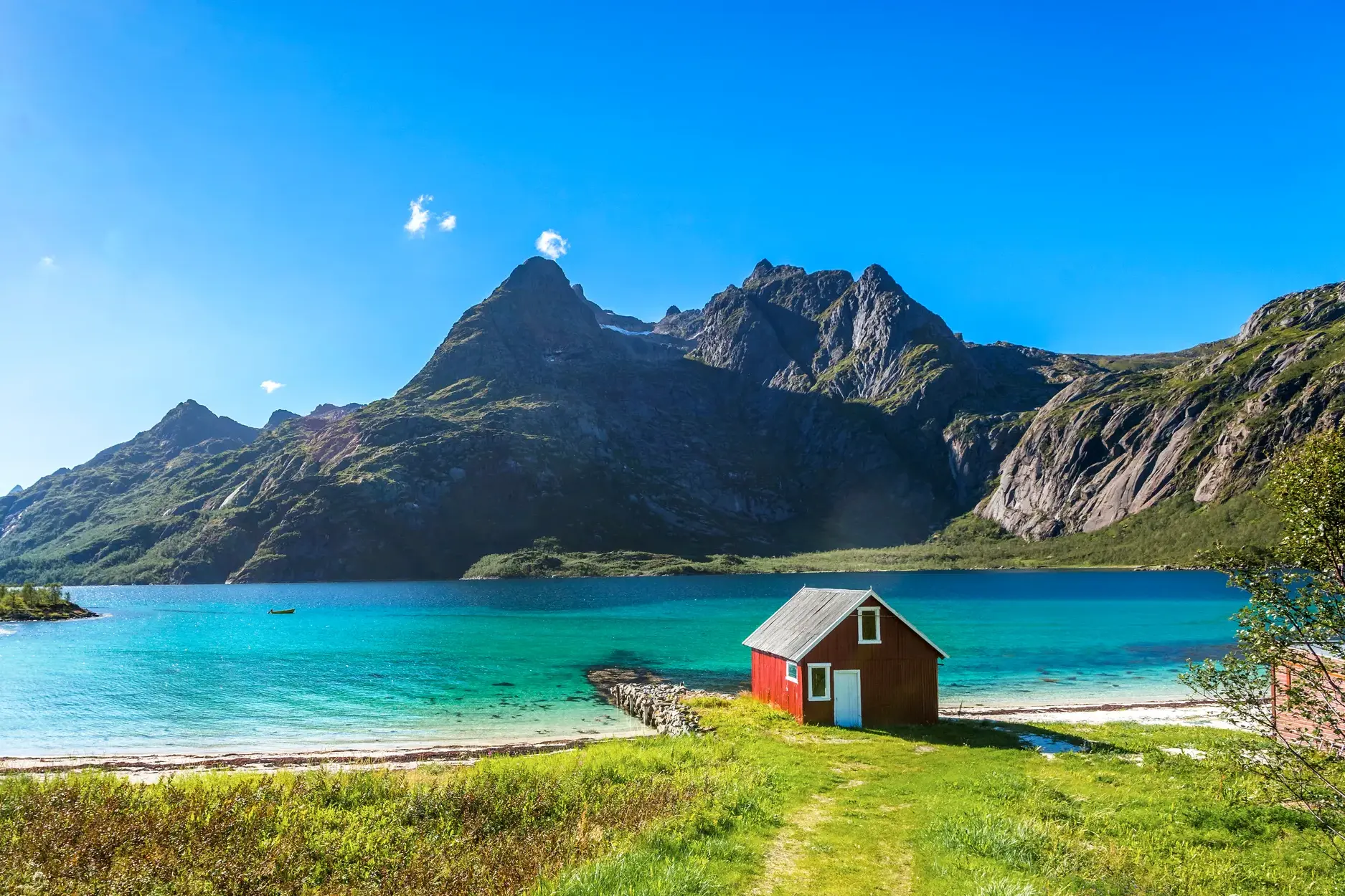 Idyllische rote Fischerhütte am Ufer eines Fjords auf den Lofoten mit türkisem Wasser und imposanter Bergkulisse.