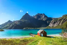 Idyllische rote Fischerhütte am Ufer eines Fjords auf den Lofoten mit türkisem Wasser und imposanter Bergkulisse.