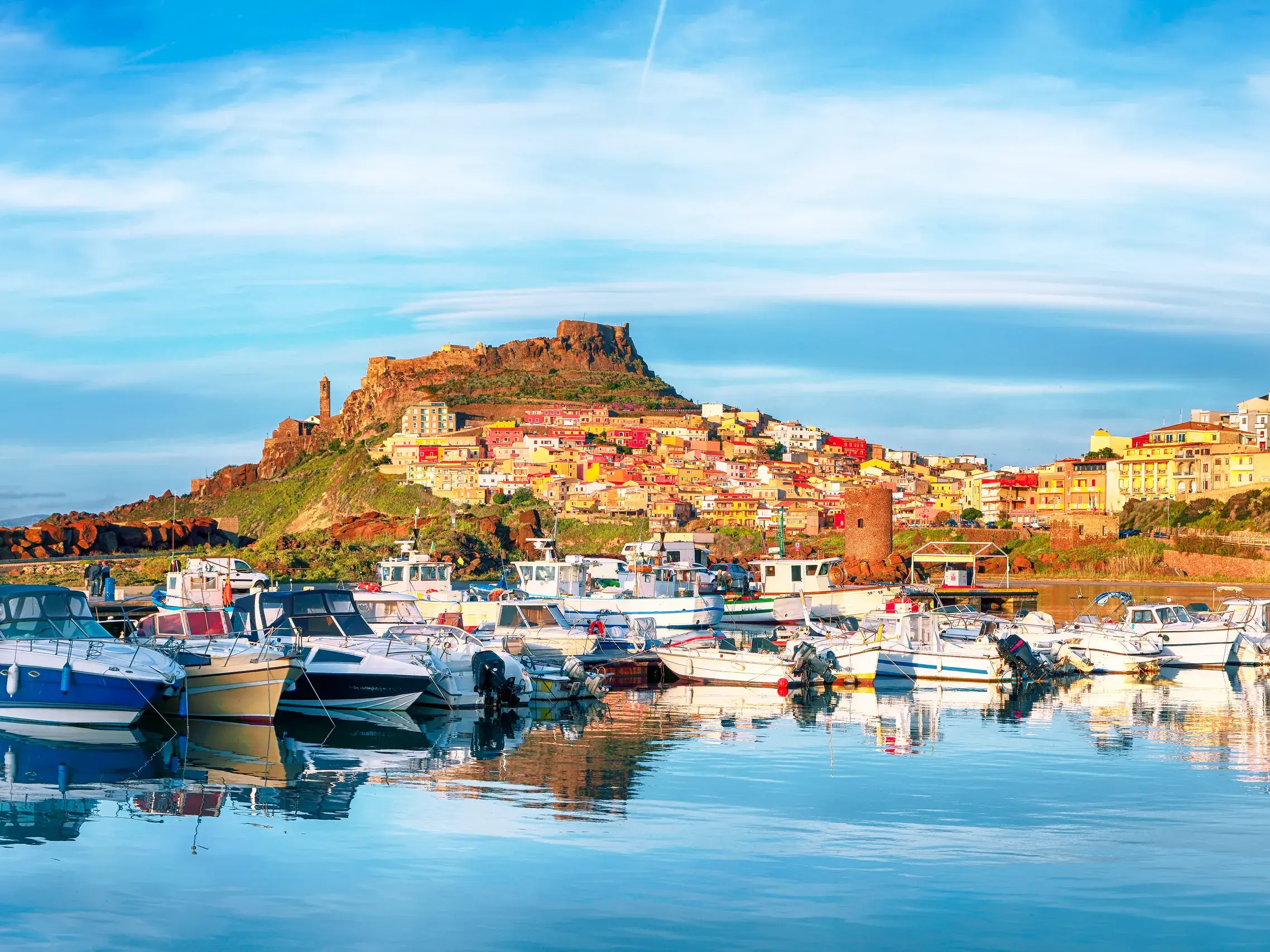 Im Hafen von Castelsardo auf Sardinien spiegeln sich Boote im ruhigen Wasser vor der malerischen, bunten Festungsstadt.