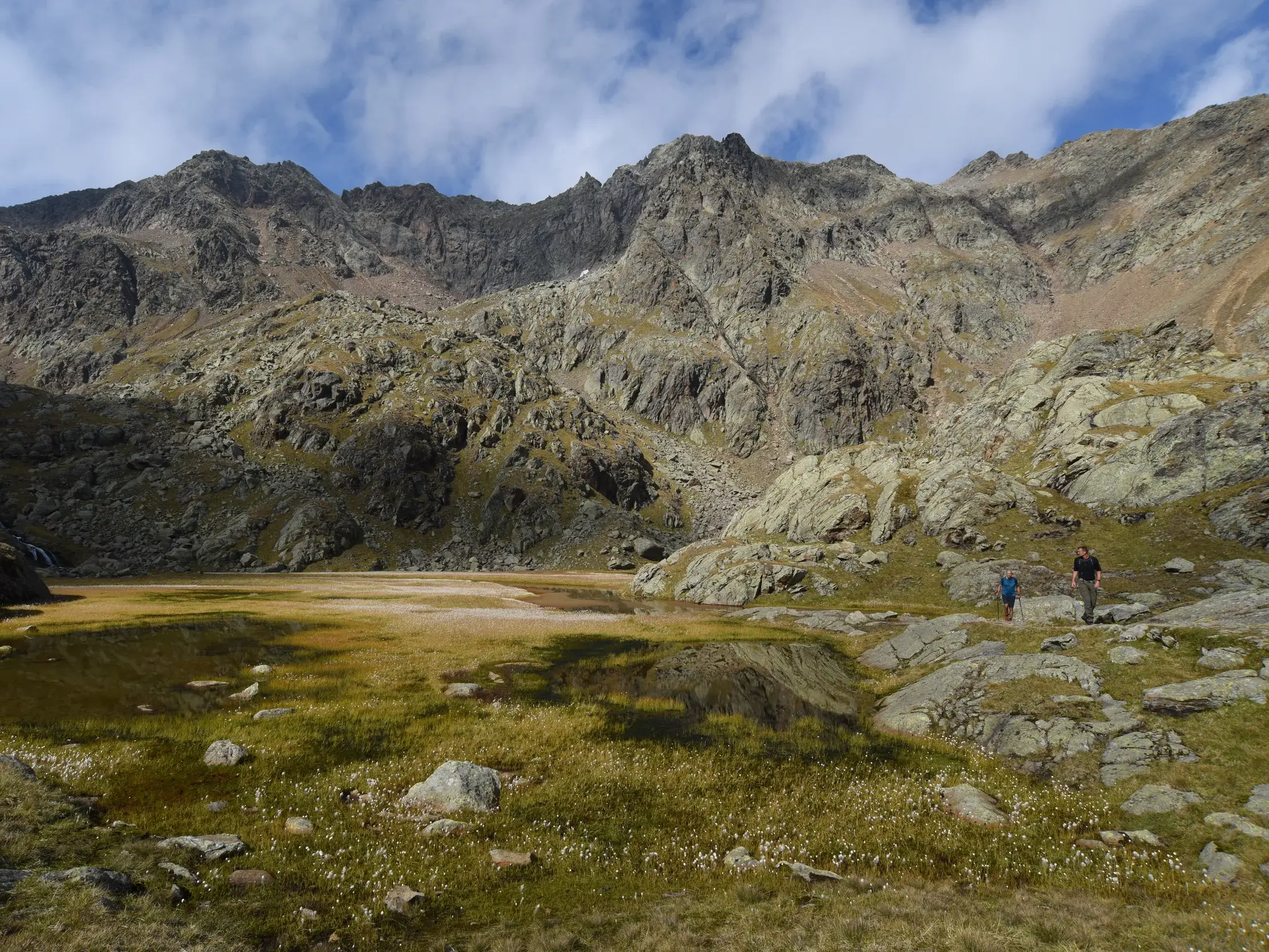 Zwei Wanderer auf einem steinigen Pfad entlang einer sumpfigen Wiese vor der Kulisse einer schroffen, felsigen Bergkette.