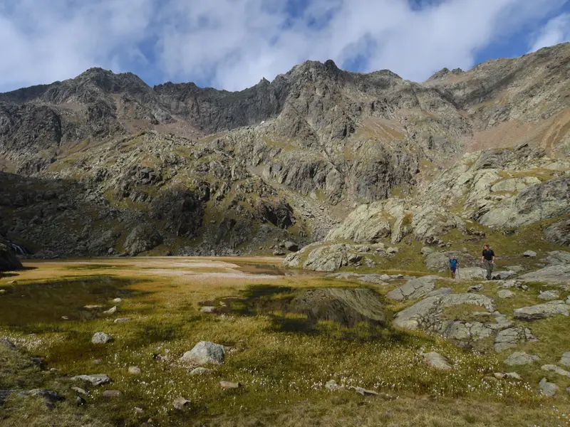 Zwei Wanderer auf einem steinigen Pfad entlang einer sumpfigen Wiese vor der Kulisse einer schroffen, felsigen Bergkette.