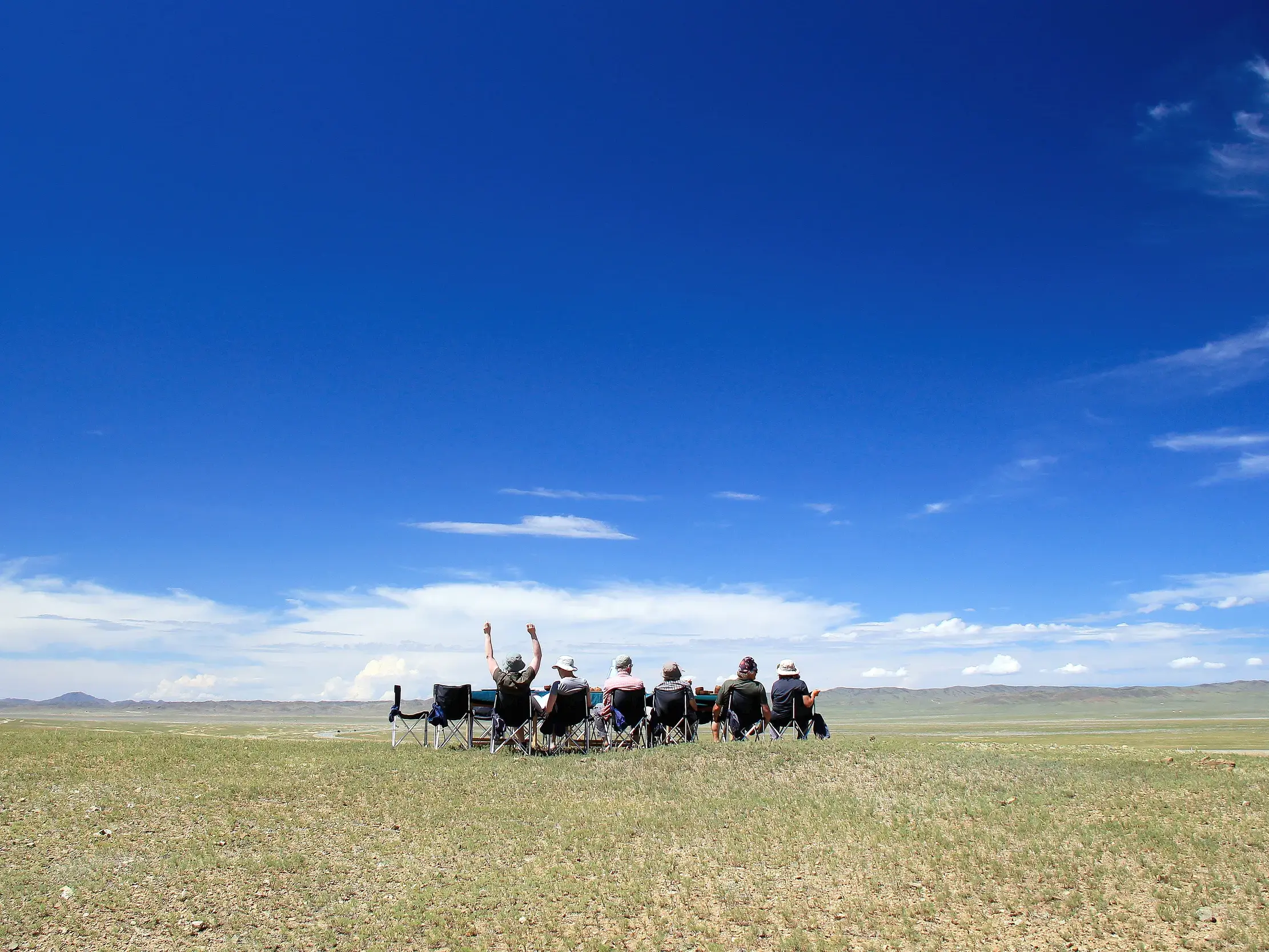 Eine kleine Reisegruppe macht eine Rast in der endlosen Weite der mongolischen Steppe, eine Person jubelt unter blauem Himmel.