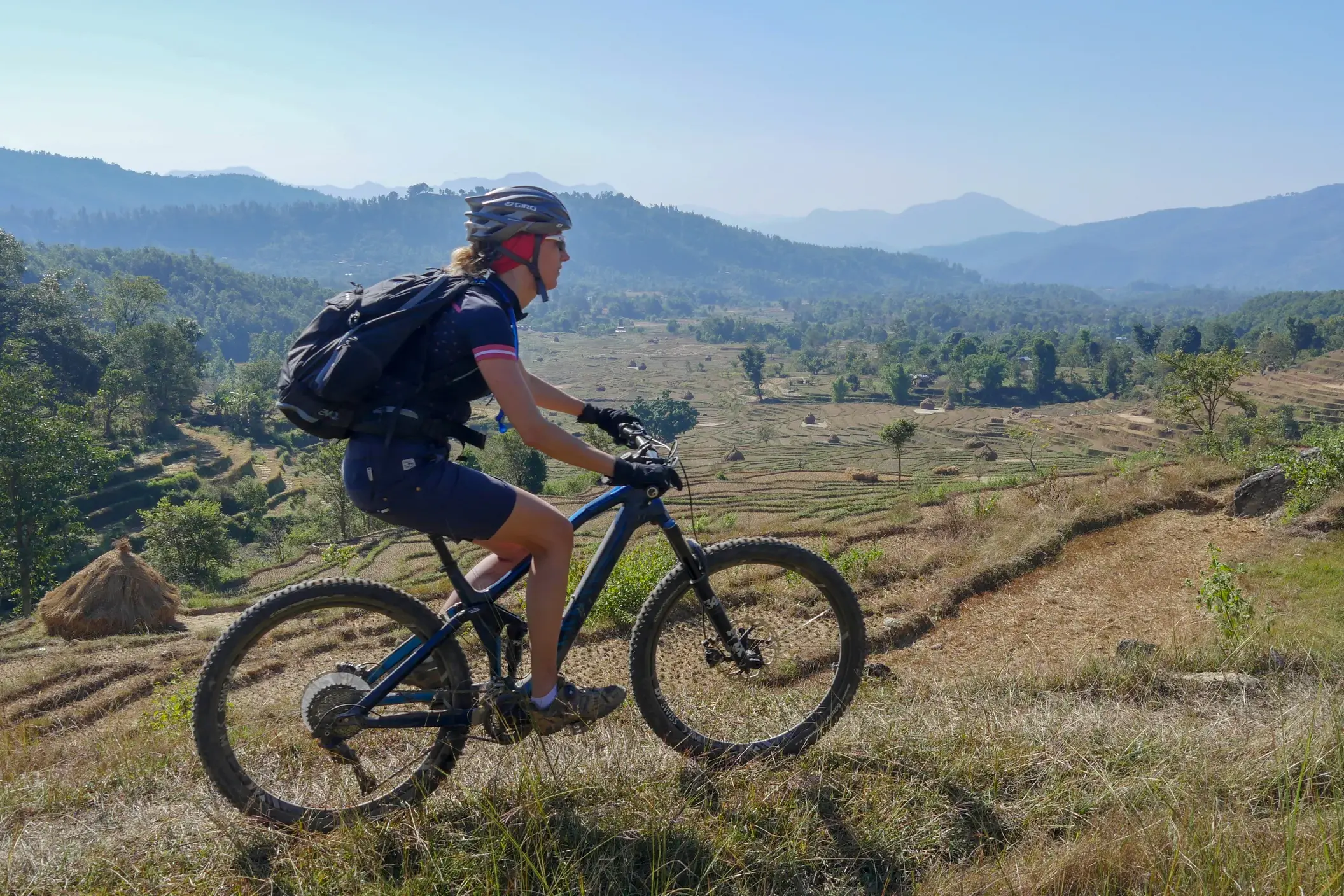 Eine Mountainbikerin auf einer Radreise in Nepal blickt von einem Trail über terrassierte Felder in die Bergwelt des Himalaya.