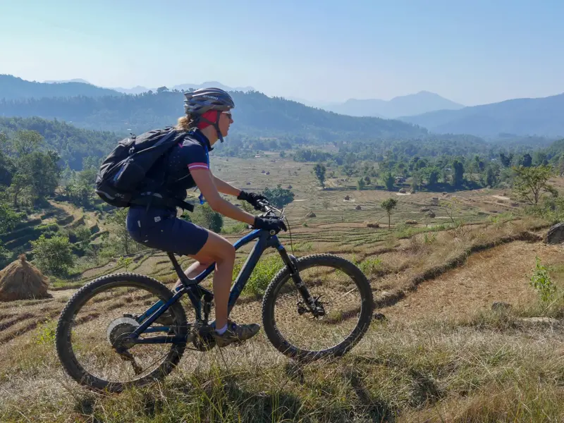 Eine Mountainbikerin auf einer Radreise in Nepal blickt von einem Trail über terrassierte Felder in die Bergwelt des Himalaya.