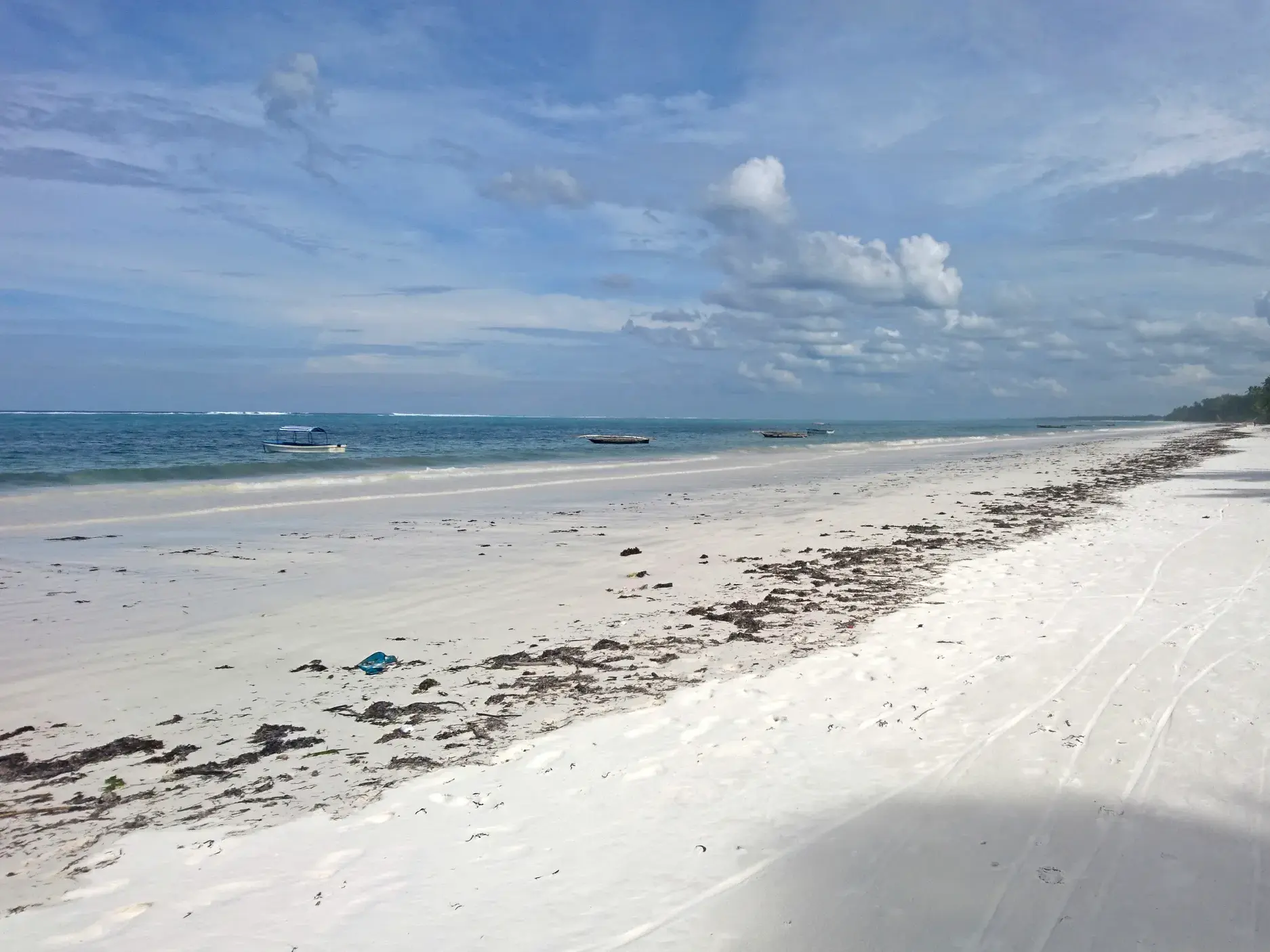 Weitläufiger, weißer Sandstrand auf Sansibar mit angespültem Seegras und kleinen Booten im türkisen Indischen Ozean.