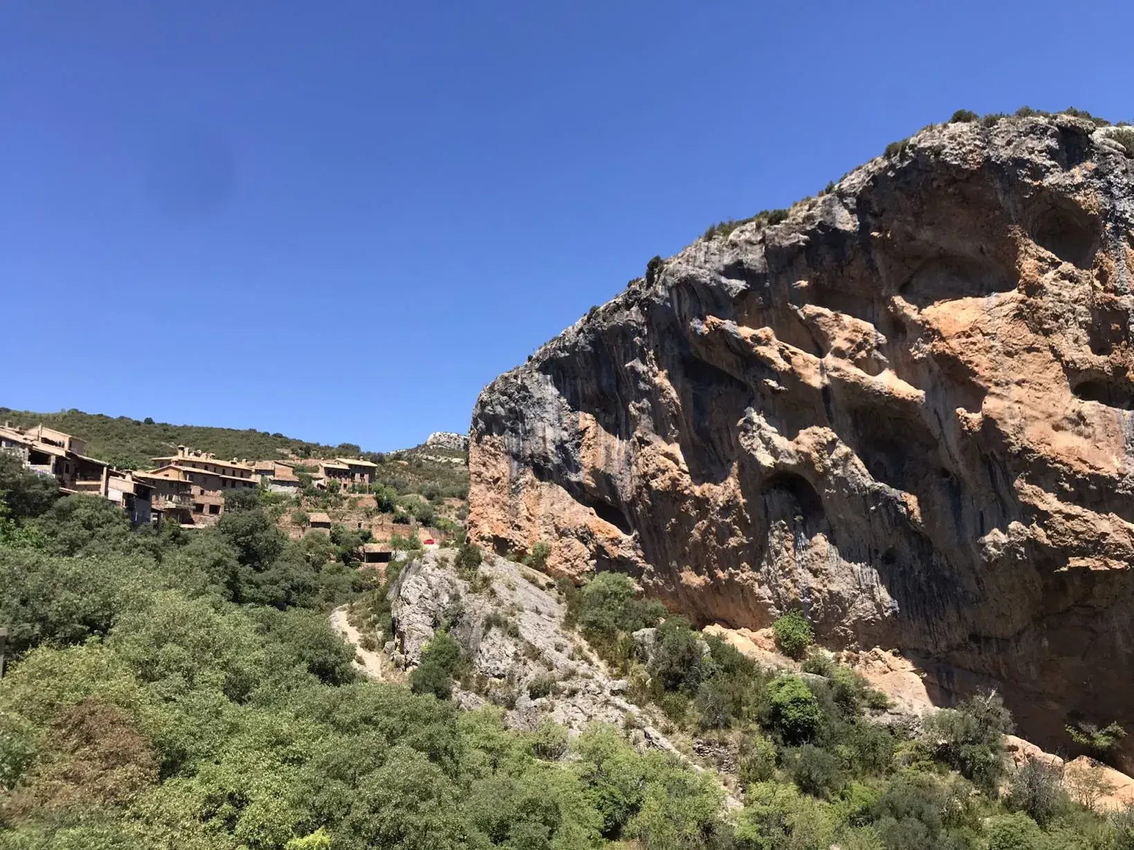 Blick auf das Bergdorf Alquézar in Aragonien, das sich an einen grünen Hang neben einer steilen Felswand schmiegt.
