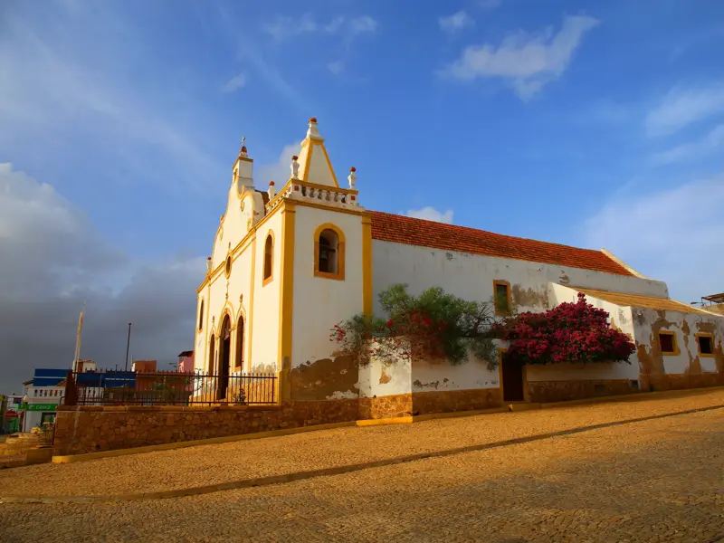 Historische Kapelle Nossa Senhora do Socorro auf der Kapverden-Insel Fogo mit weiß-gelber Fassade an einer Gasse aus Kopfsteinpflaster.