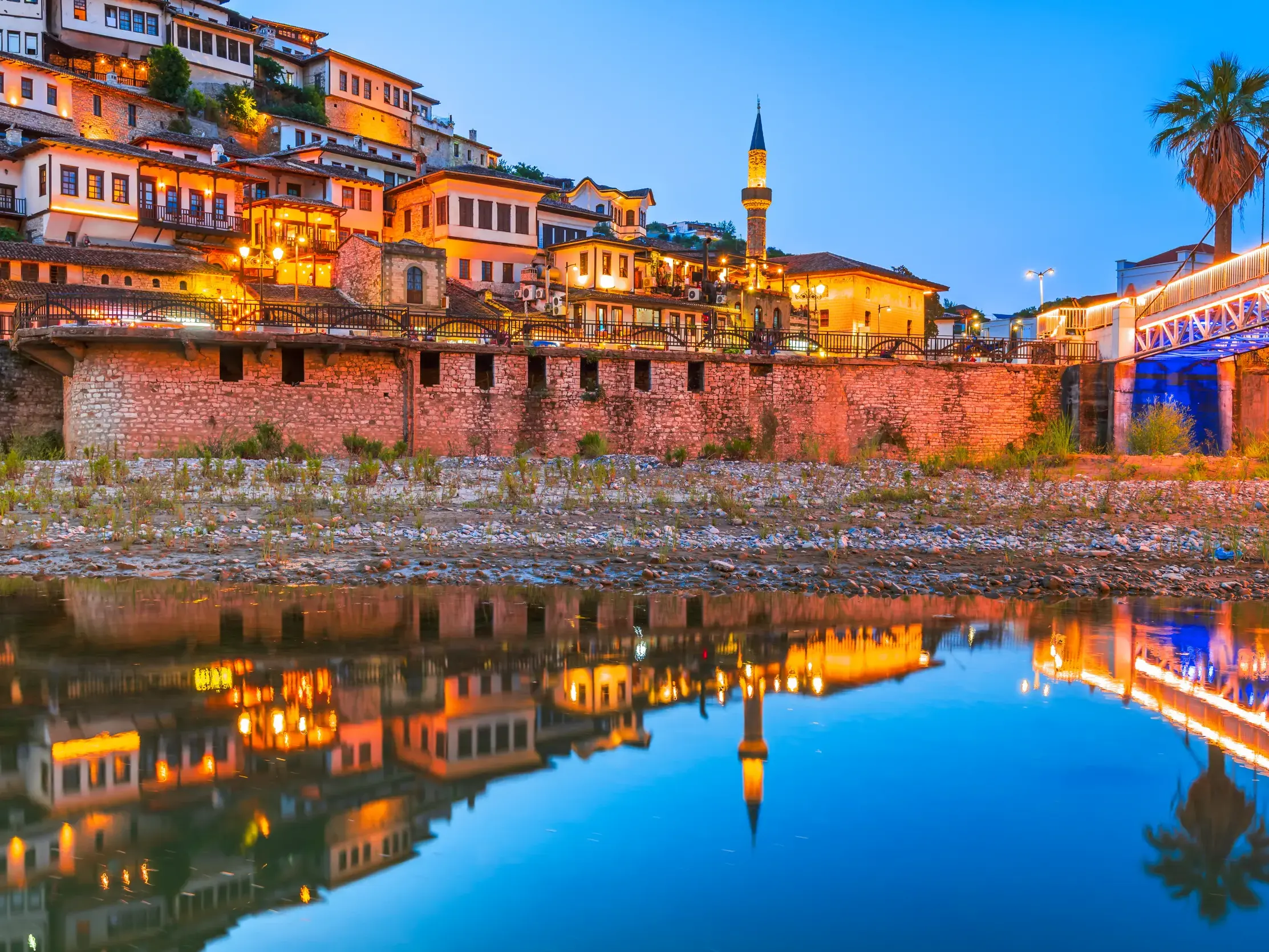 Die beleuchteten osmanischen Häuser der UNESCO-Stadt Berat spiegeln sich bei Dämmerung im ruhigen Wasser des Flusses Osumi.