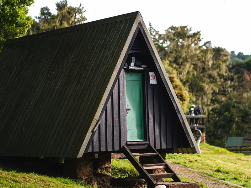 Rustikale Berghütte mit grüner Tür im Camp der Marangu-Route am Kilimanjaro, umgeben von üppiger Vegetation.