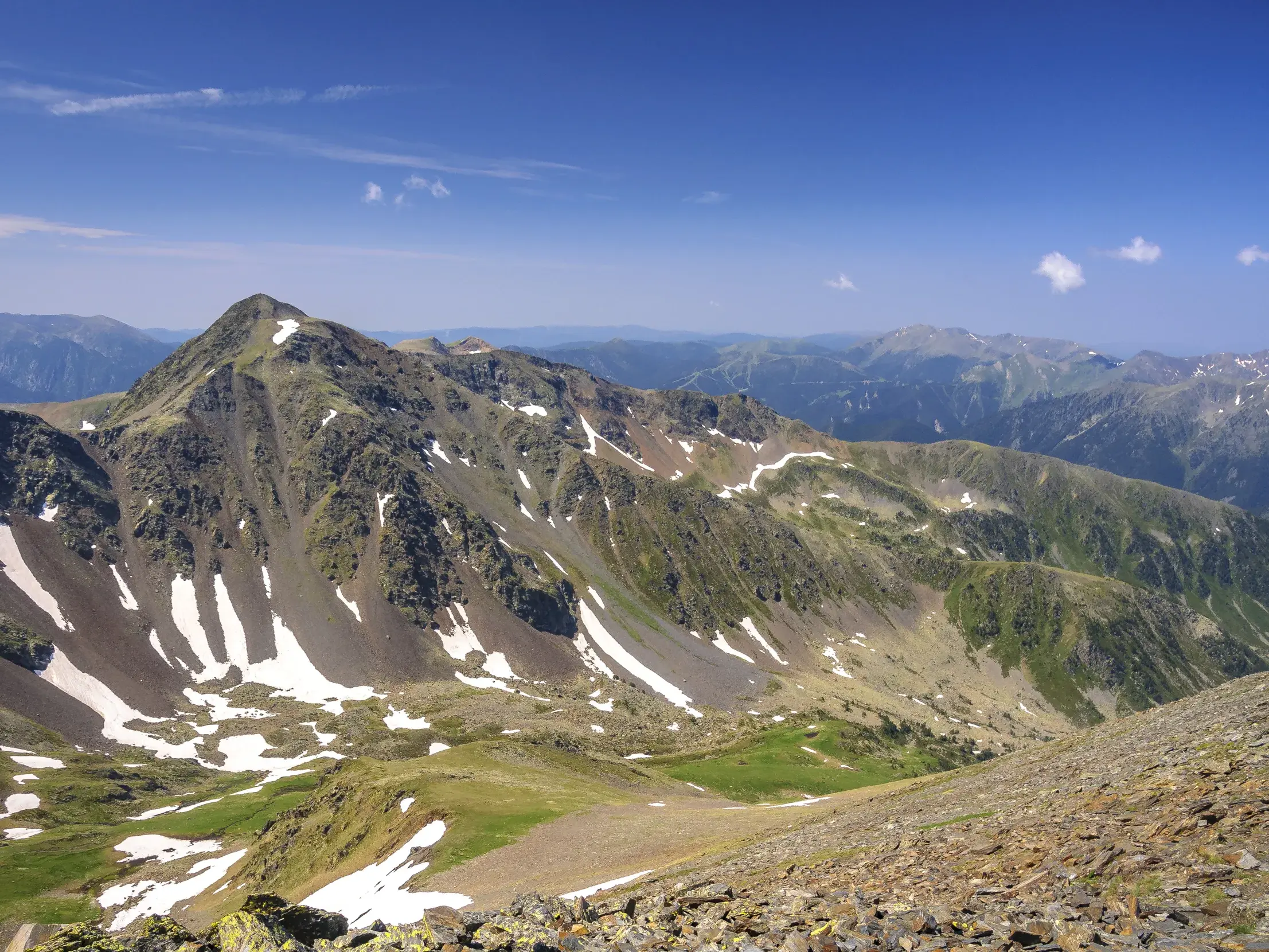 Felsiger Gipfel des Pic de Montmalús in Andorra, umgeben von grünen Tälern mit Schneeresten an einem sonnigen Wandertag.