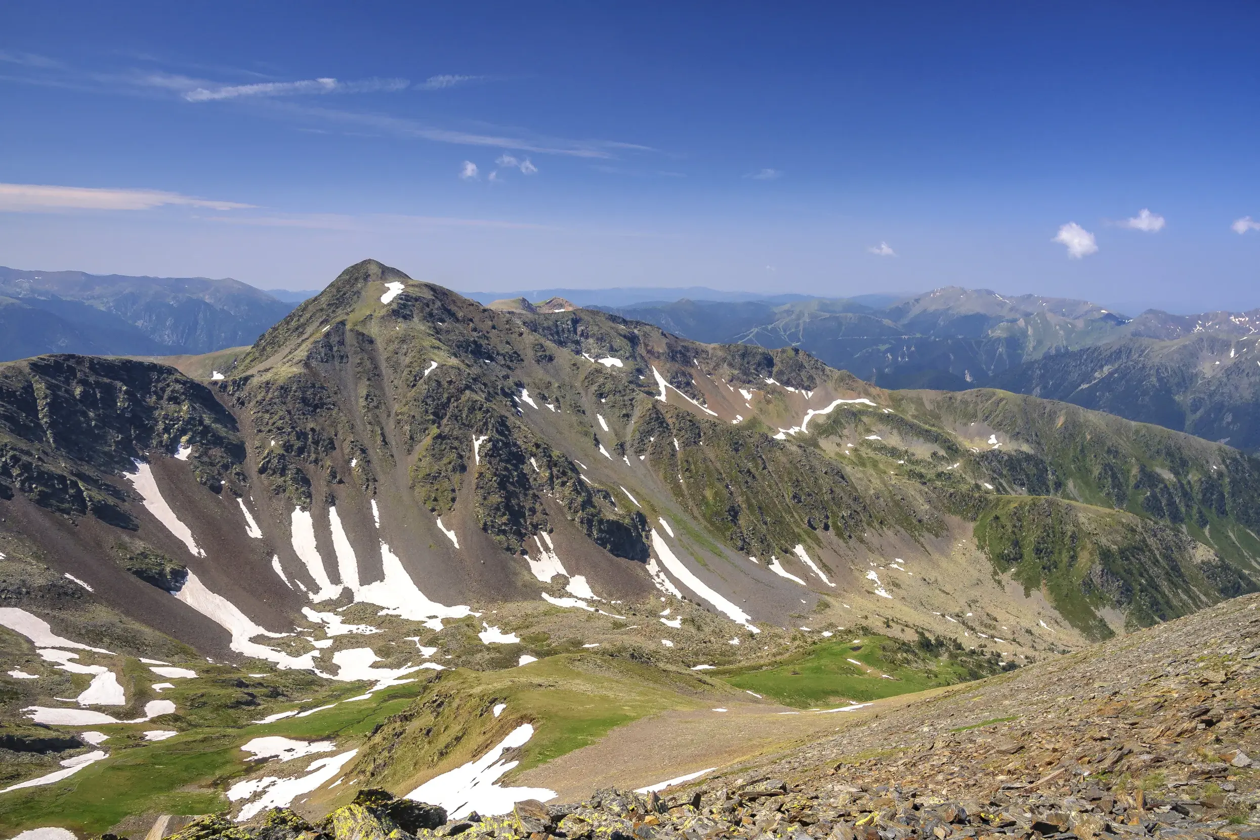 Felsiger Gipfel des Pic de Montmalús in Andorra, umgeben von grünen Tälern mit Schneeresten an einem sonnigen Wandertag.