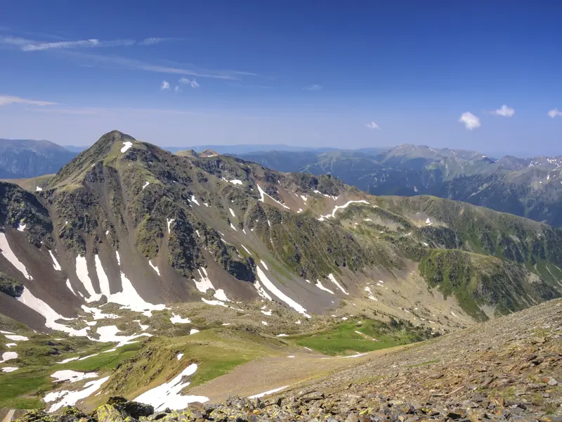 Felsiger Gipfel des Pic de Montmalús in Andorra, umgeben von grünen Tälern mit Schneeresten an einem sonnigen Wandertag.