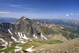 Felsiger Gipfel des Pic de Montmalús in Andorra, umgeben von grünen Tälern mit Schneeresten an einem sonnigen Wandertag.