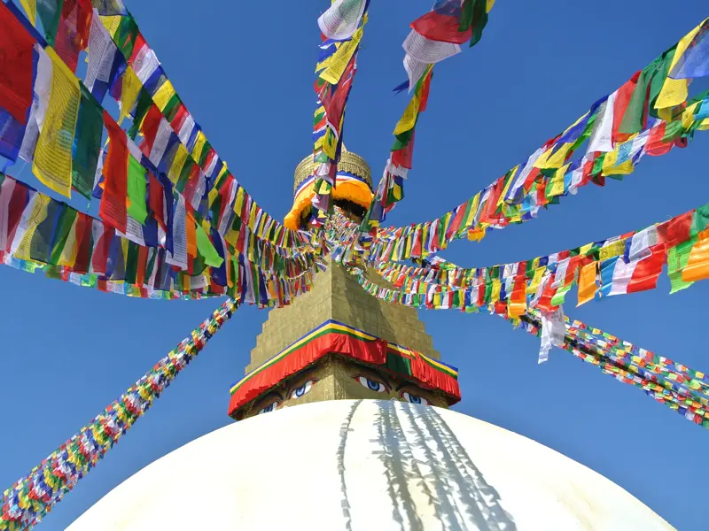 Farbenfrohe Gebetsfahnen wehen von der goldenen Spitze des Bodnath Stupa in Kathmandu, über den wachsamen Augen Buddhas.
