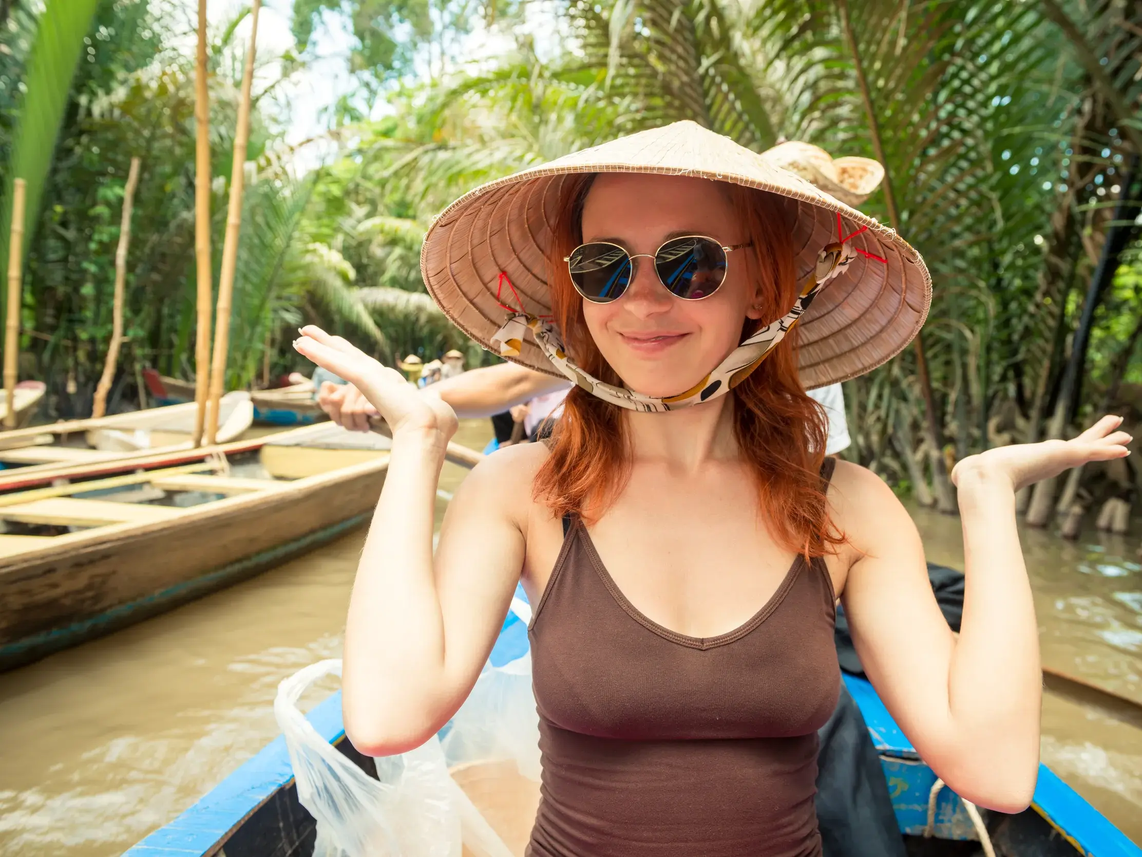 Eine junge Frau mit traditionellem Reishut und Sonnenbrille genießt eine Bootsfahrt durch die Wasserwege des Mekongdeltas in Vietnam.