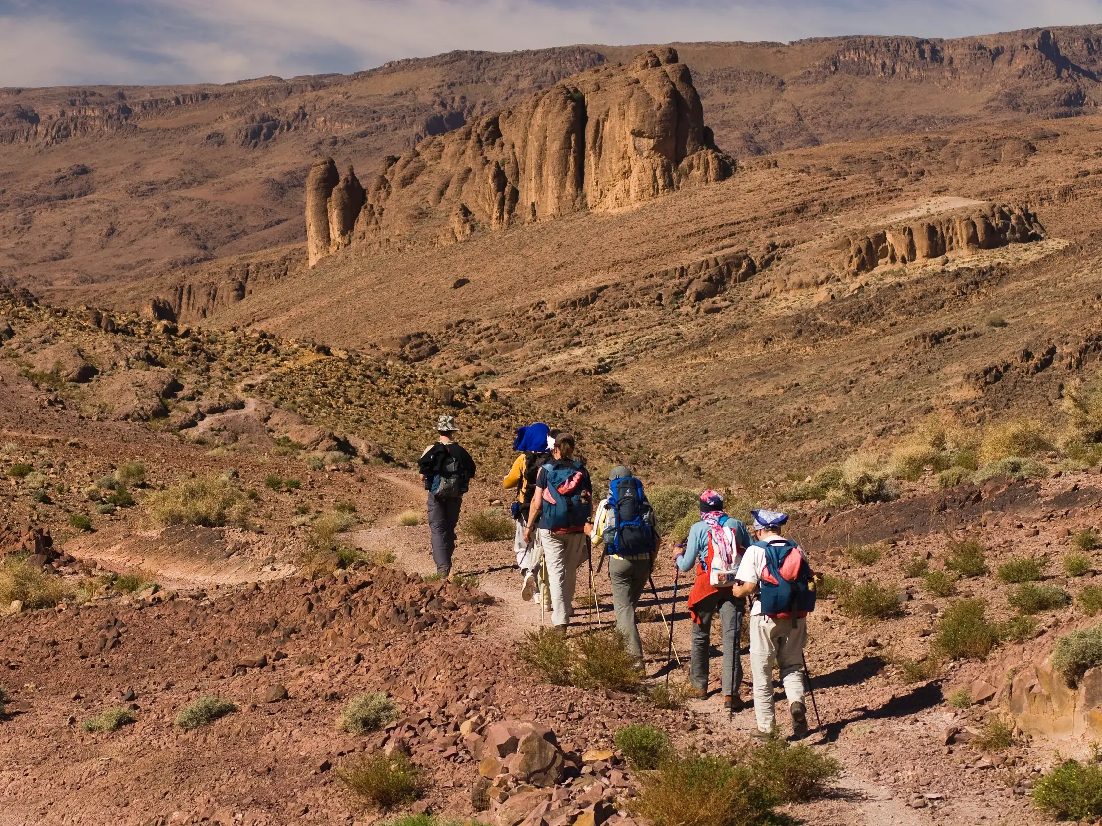 Eine Wandergruppe durchquert auf einem schmalen Pfad die rote, zerklüftete Vulkanlandschaft des Djebel Saghro Gebirges in Marokko.