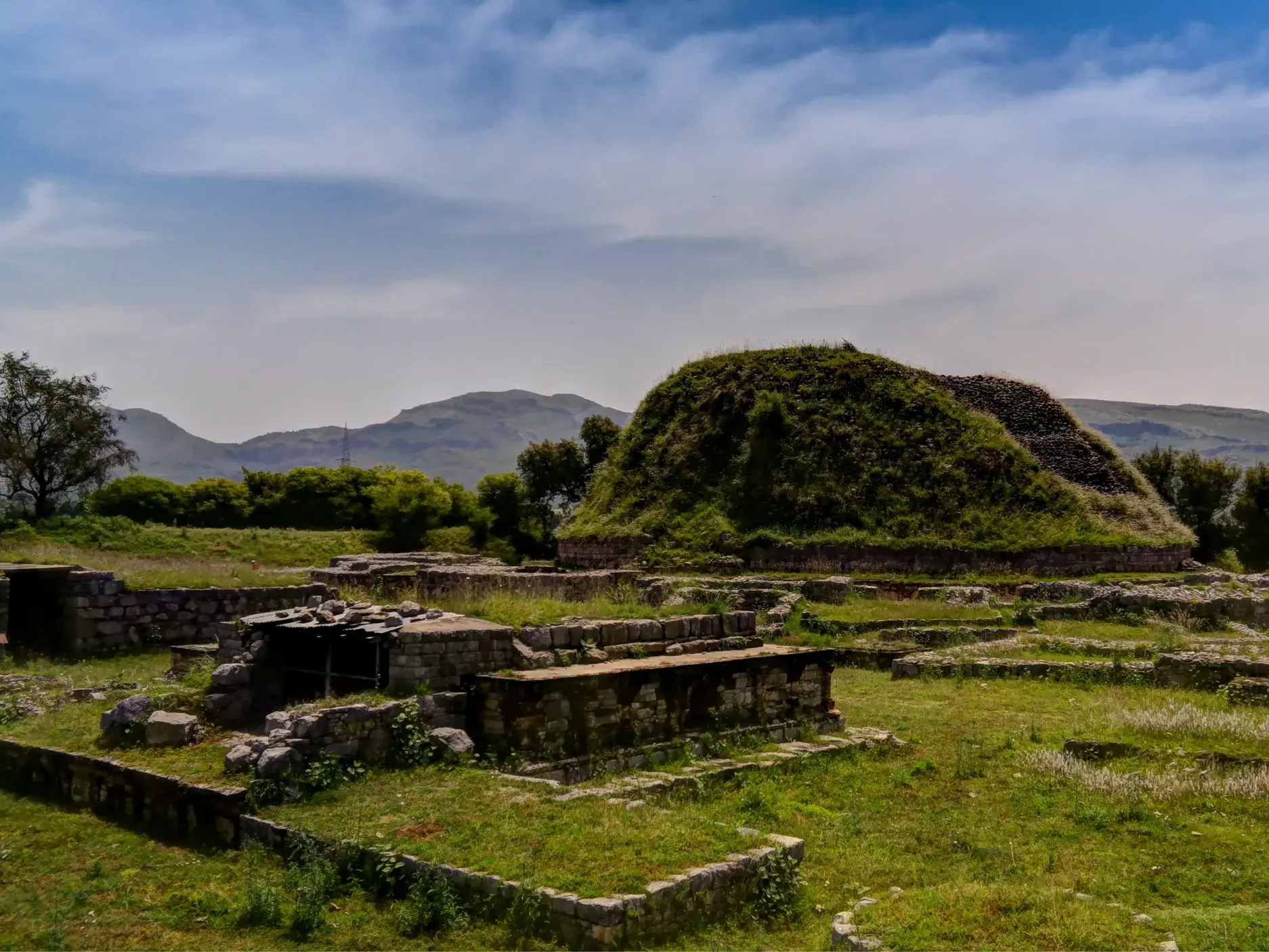 Die antiken, von Gras bewachsenen Ruinen von Taxila in Pakistan mit der Dharmarajika-Stupa im Hintergrund vor Bergkulisse.