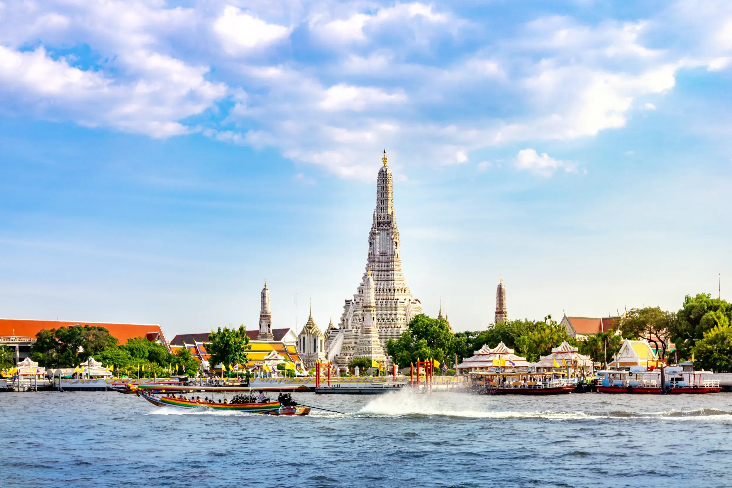 Ein Longtail-Boot mit Passagieren überquert den Chao Phraya Fluss vor dem Wahrzeichen Wat Arun in Bangkok an einem sonnigen Tag.