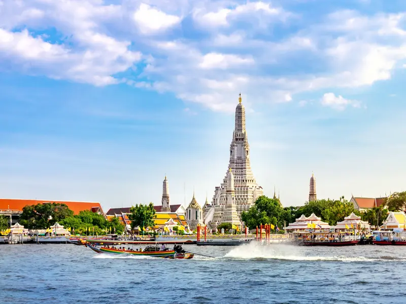 Ein Longtail-Boot mit Passagieren überquert den Chao Phraya Fluss vor dem Wahrzeichen Wat Arun in Bangkok an einem sonnigen Tag.