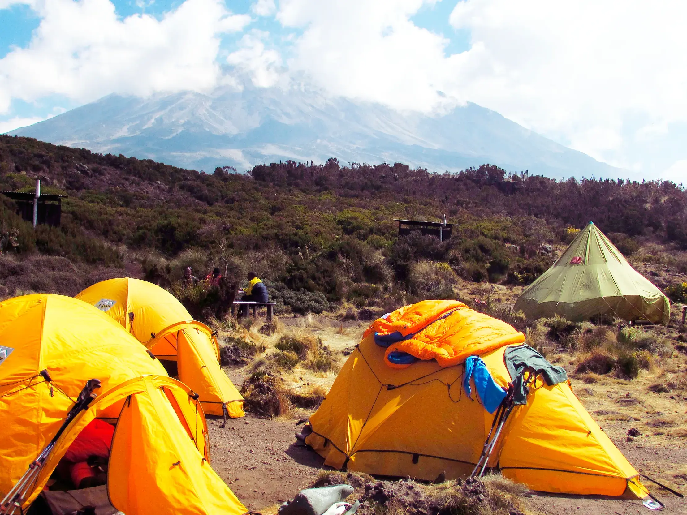 Ein Trekking-Zeltlager mit gelben Zelten liegt am Hang des Kilimanjaro, dessen majestätischer Gipfel von Wolken umspielt wird.