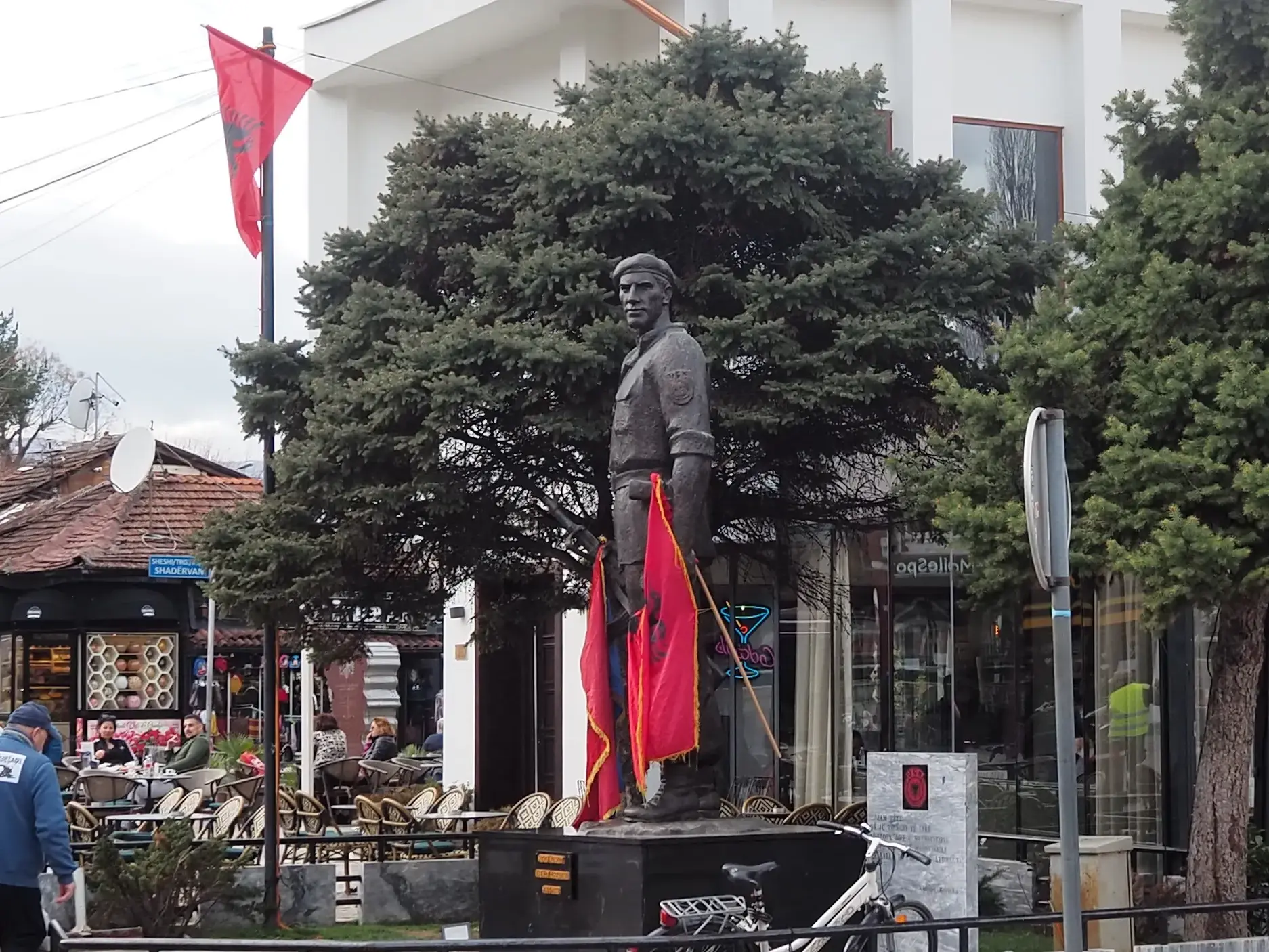 Bronzestatue eines Uniformierten mit albanischen Flaggen auf einem belebten Platz in der Kulturstadt Prizren im Kosovo.