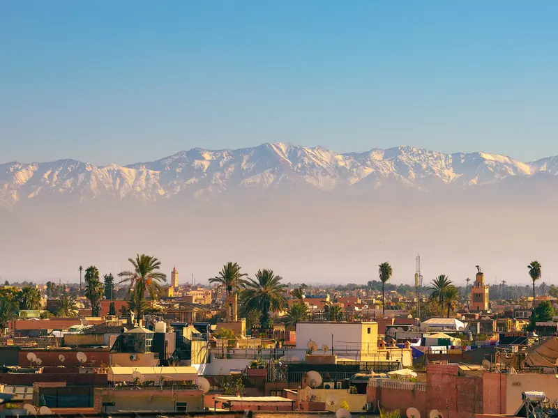 Blick von den Dächern Marrakeschs über die Medina auf die imposante, schneebedeckte Gipfelkette des Hohen Atlas in Marokko.