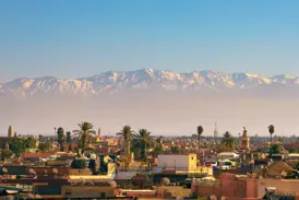 Blick von den Dächern Marrakeschs über die Medina auf die imposante, schneebedeckte Gipfelkette des Hohen Atlas in Marokko.