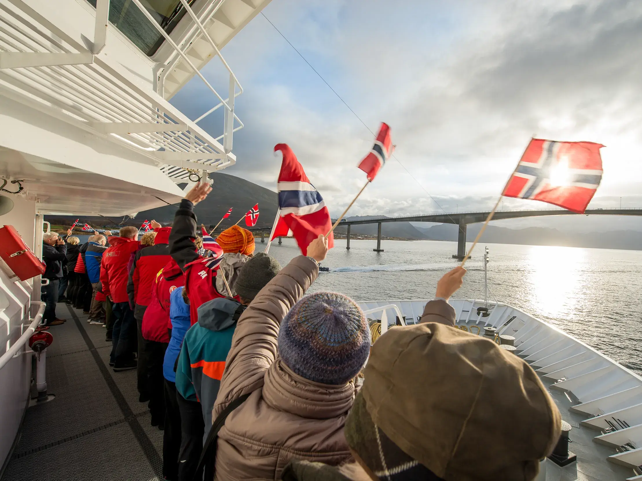 Reisegruppe winkt mit norwegischen Flaggen vom Deck eines Postschiffes entlang der Küste Norwegens bei Sonnenuntergang.