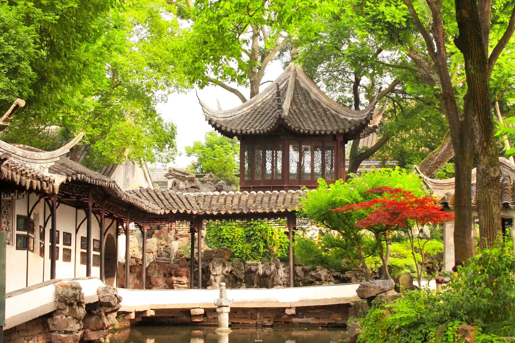 Blick auf einen hölzernen Pavillon mit Ziegeldach inmitten üppiger, grüner Bäume in einem klassischen Garten in Suzhou.