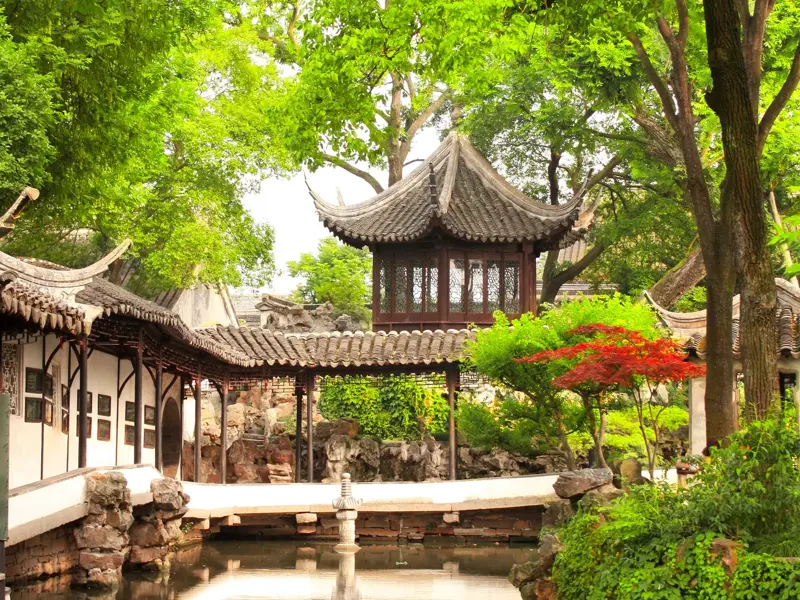 Blick auf einen hölzernen Pavillon mit Ziegeldach inmitten üppiger, grüner Bäume in einem klassischen Garten in Suzhou.