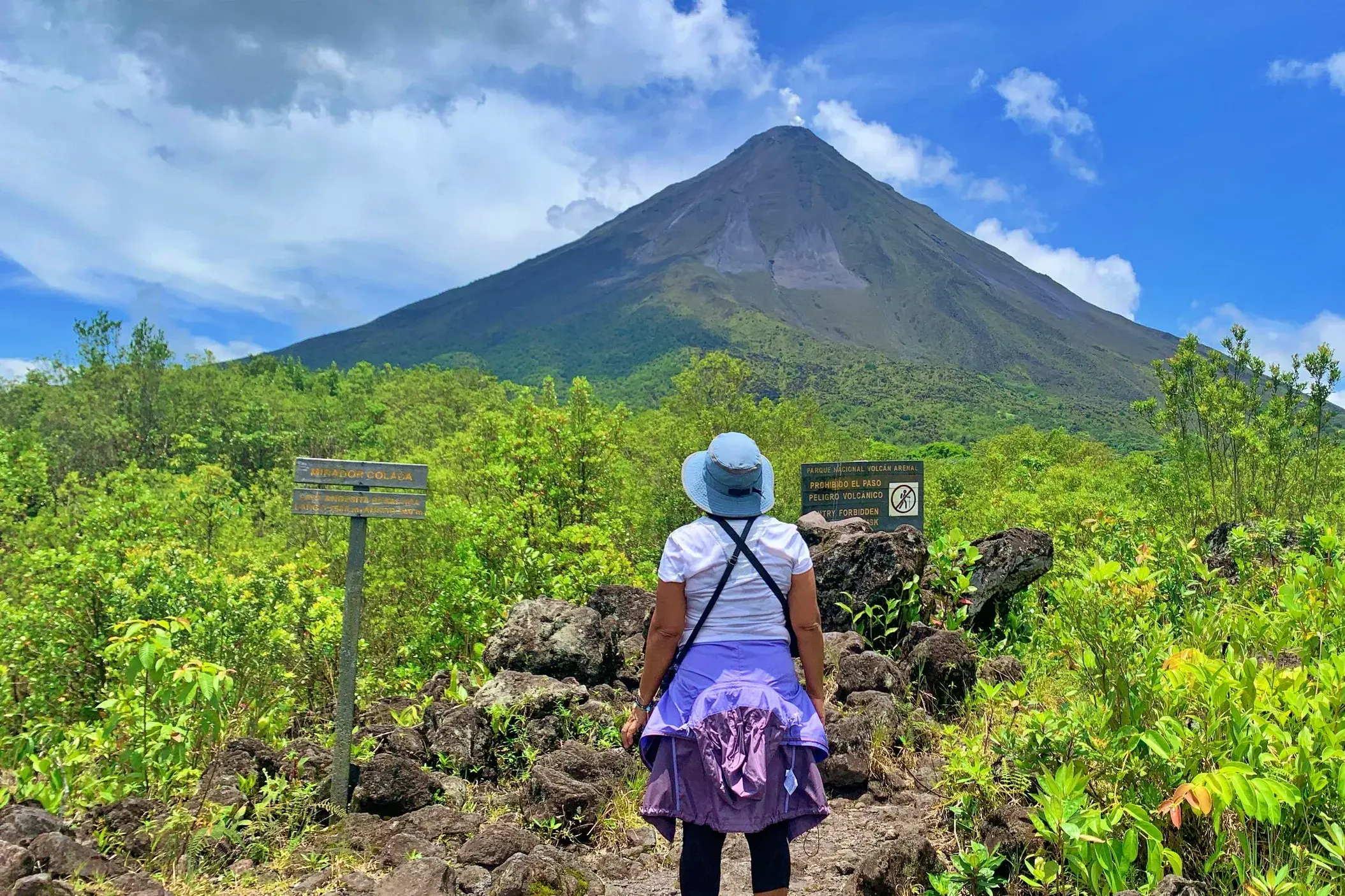 Eine Wanderin auf einem Pfad aus Lavagestein blickt auf den von tropischem Grün umgebenen Vulkan Arenal in Costa Rica.