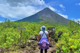 Eine Wanderin auf einem Pfad aus Lavagestein blickt auf den von tropischem Grün umgebenen Vulkan Arenal in Costa Rica.