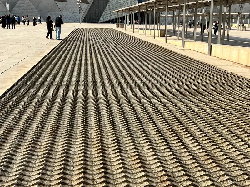Gezacktes Steinmuster auf dem Vorplatz des Grand Egyptian Museum in Kairo mit Besuchern vor der modernen Fassade.
