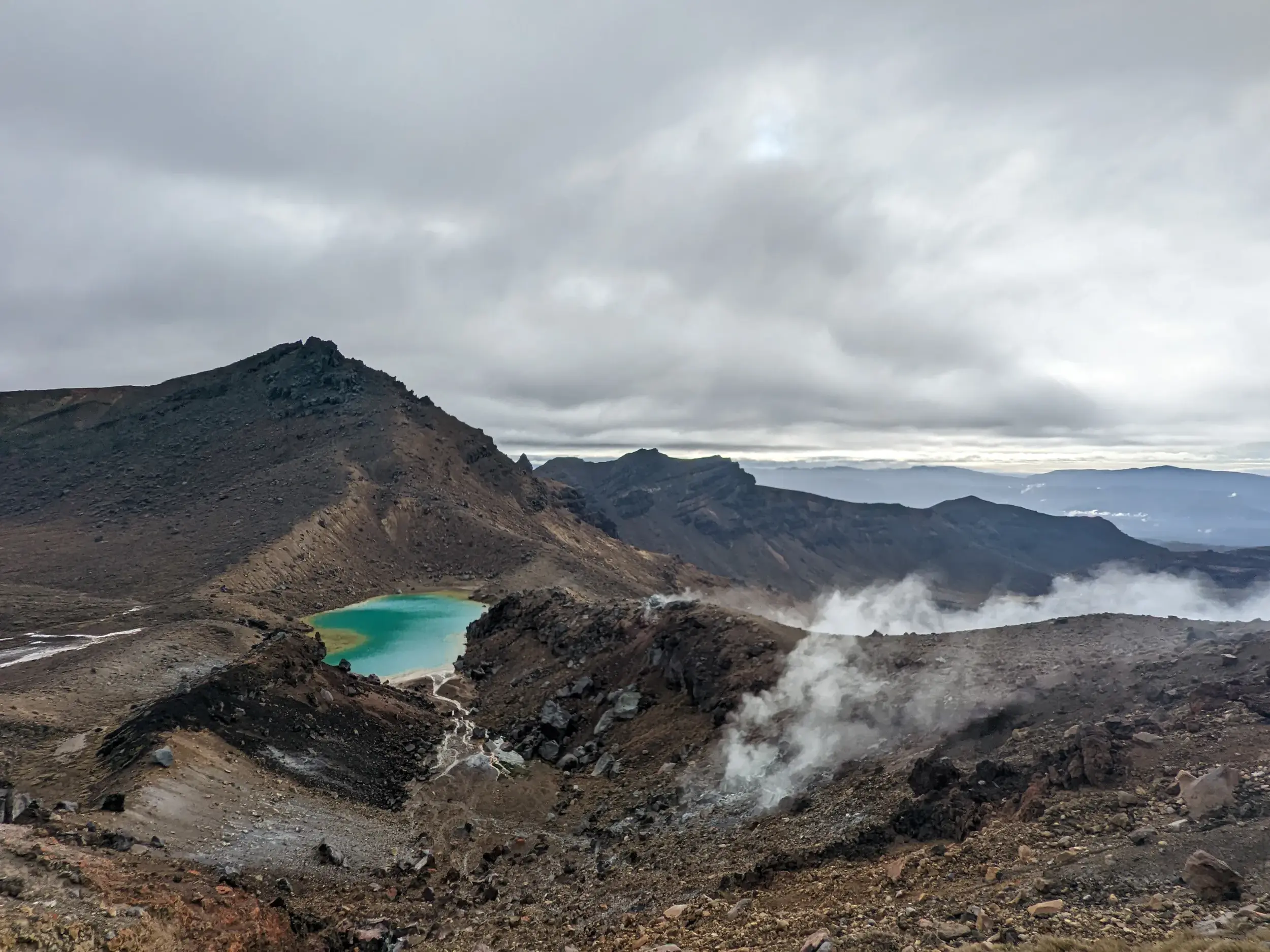 Rauhe Vulkanlandschaft auf dem Tongariro Crossing mit dampfenden Fumarolen und einem der leuchtend smaragdgrünen Seen.