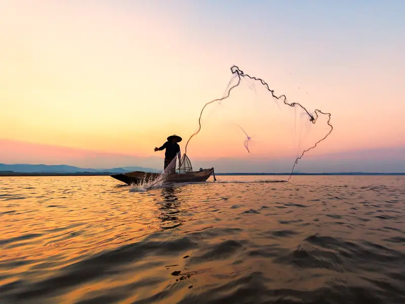 Fischer mit traditionellem Kegelhut wirft sein Netz von einem Boot auf dem Mekong bei malerischem Sonnenuntergang in Südostasien.