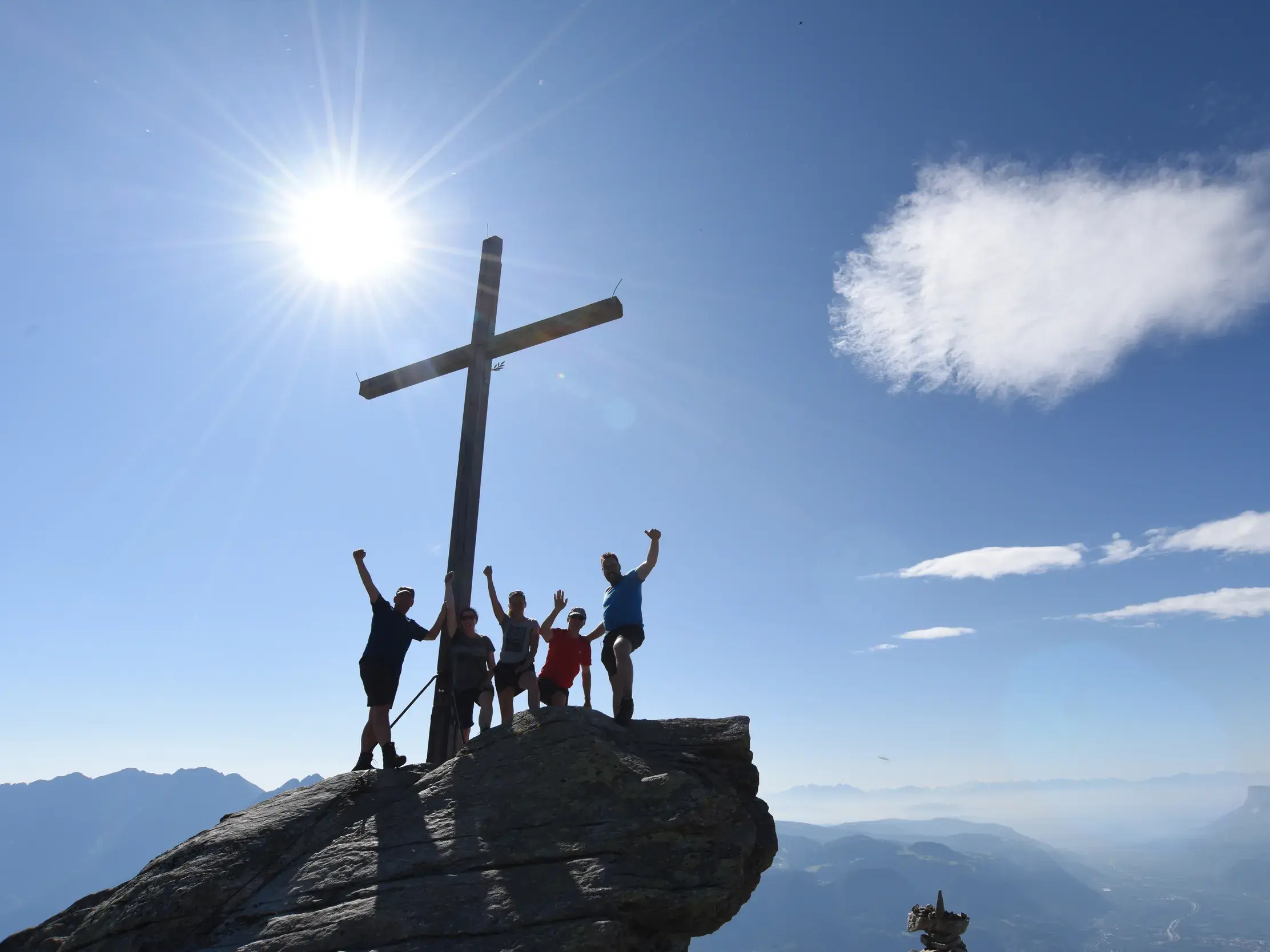 Wandergruppe jubelt am Gipfelkreuz der Mutspitze mit Panoramablick über die Texelgruppe in Südtirol bei Sonnenschein.