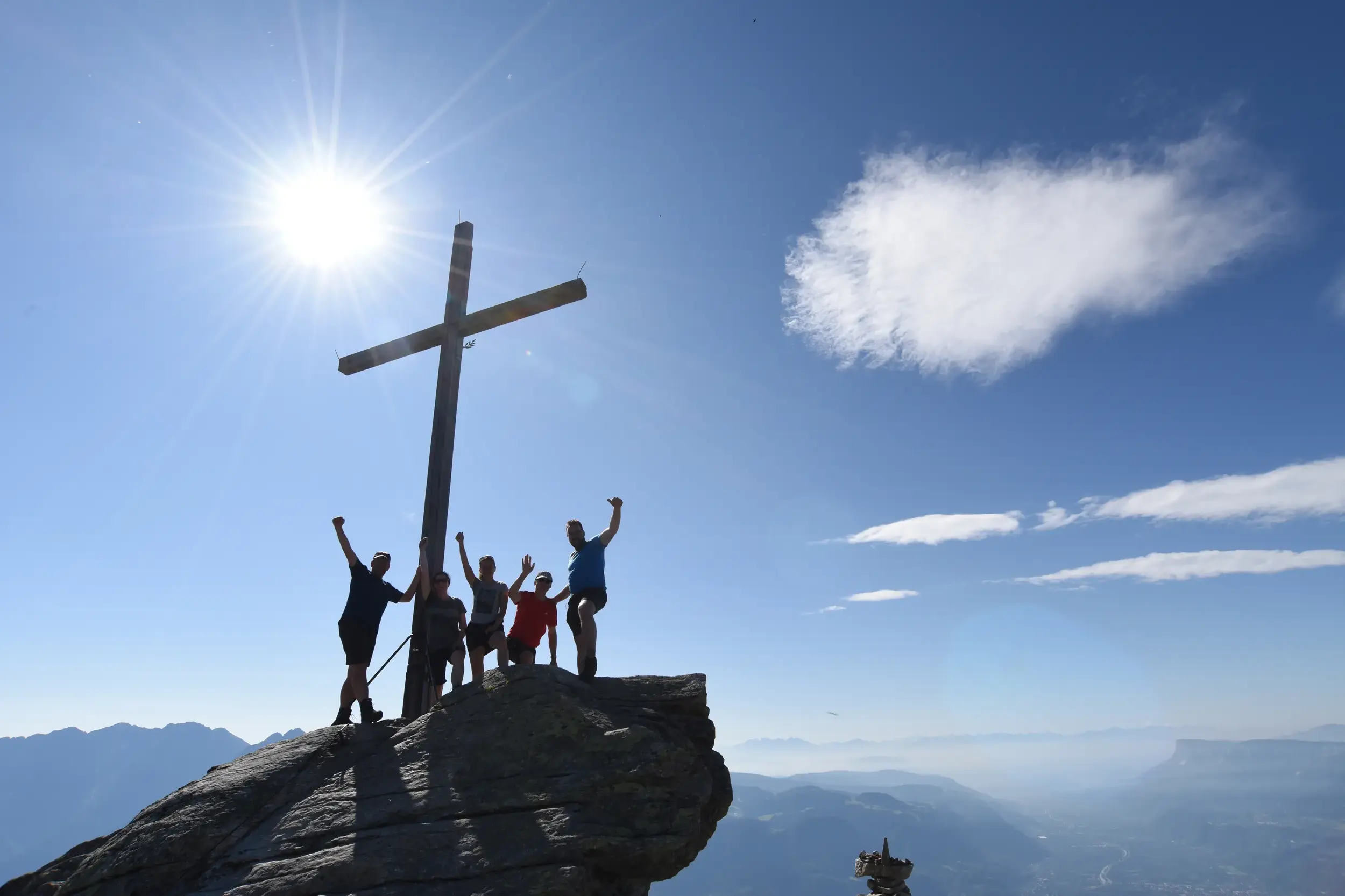 Wandergruppe jubelt am Gipfelkreuz der Mutspitze mit Panoramablick über die Texelgruppe in Südtirol bei Sonnenschein.