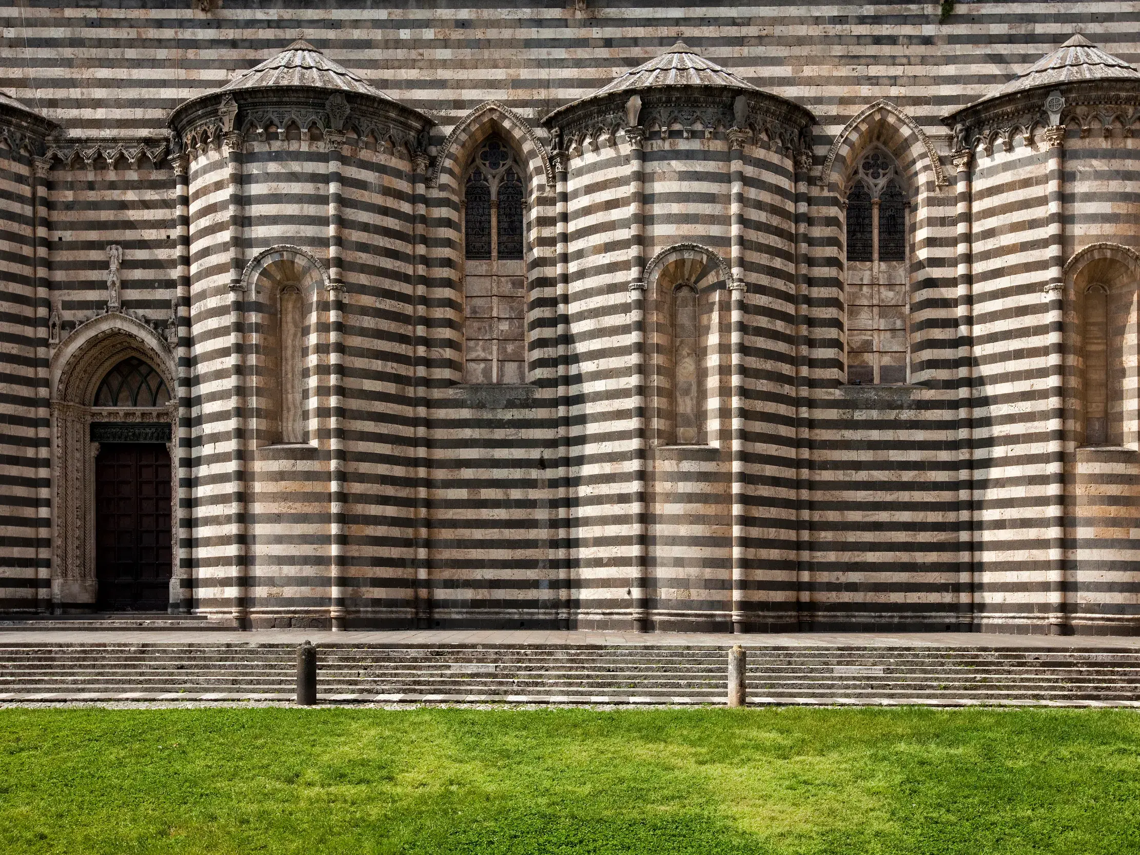 Die beeindruckende, schwarz-weiß gestreifte Fassade des gotischen Doms von Orvieto in Umbrien, davor eine grüne Rasenfläche.