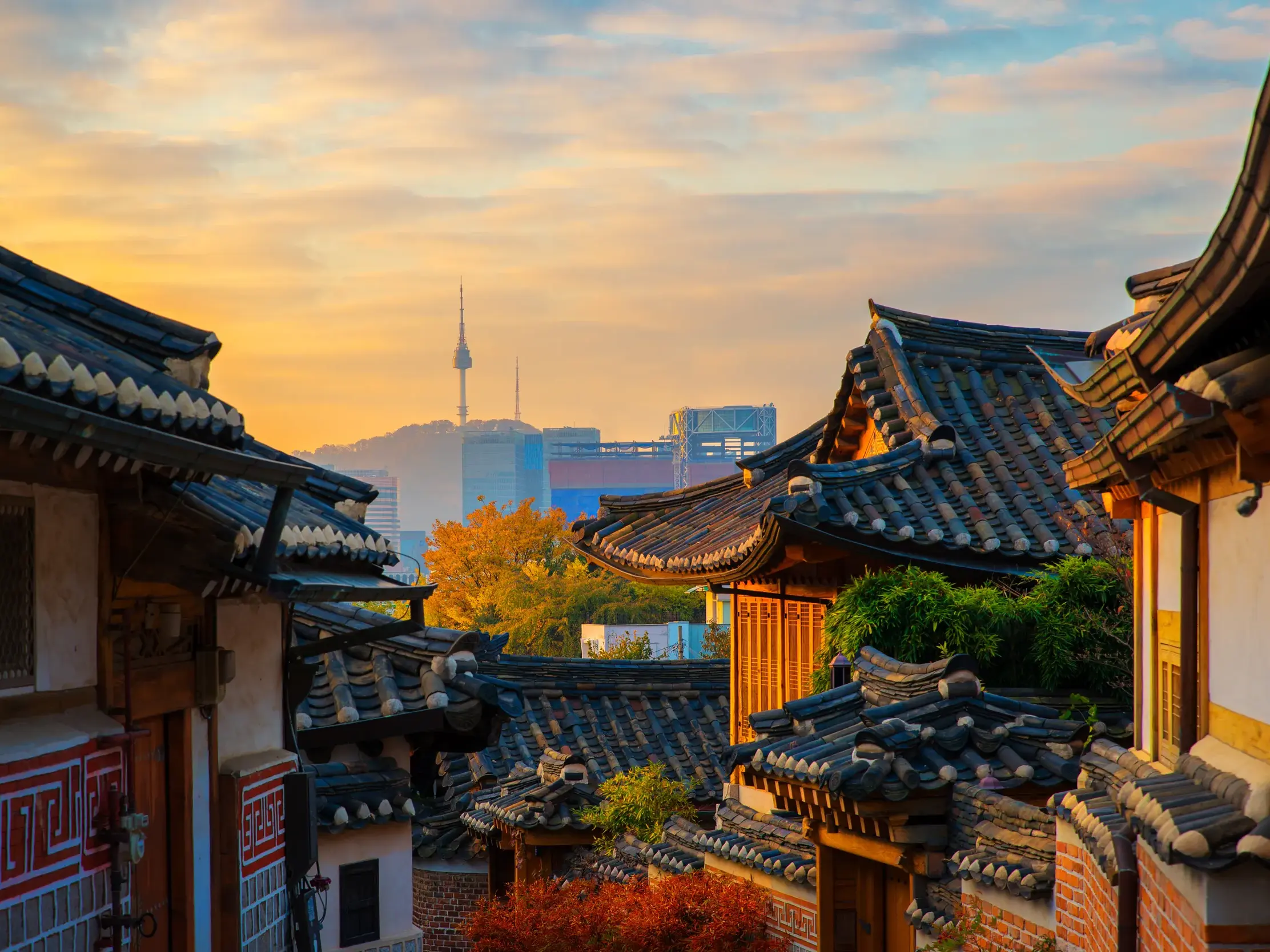 Traditionelle Hanok-Dächer in einem historischen Viertel Seouls vor der modernen Skyline mit dem N Seoul Tower bei Sonnenuntergang.