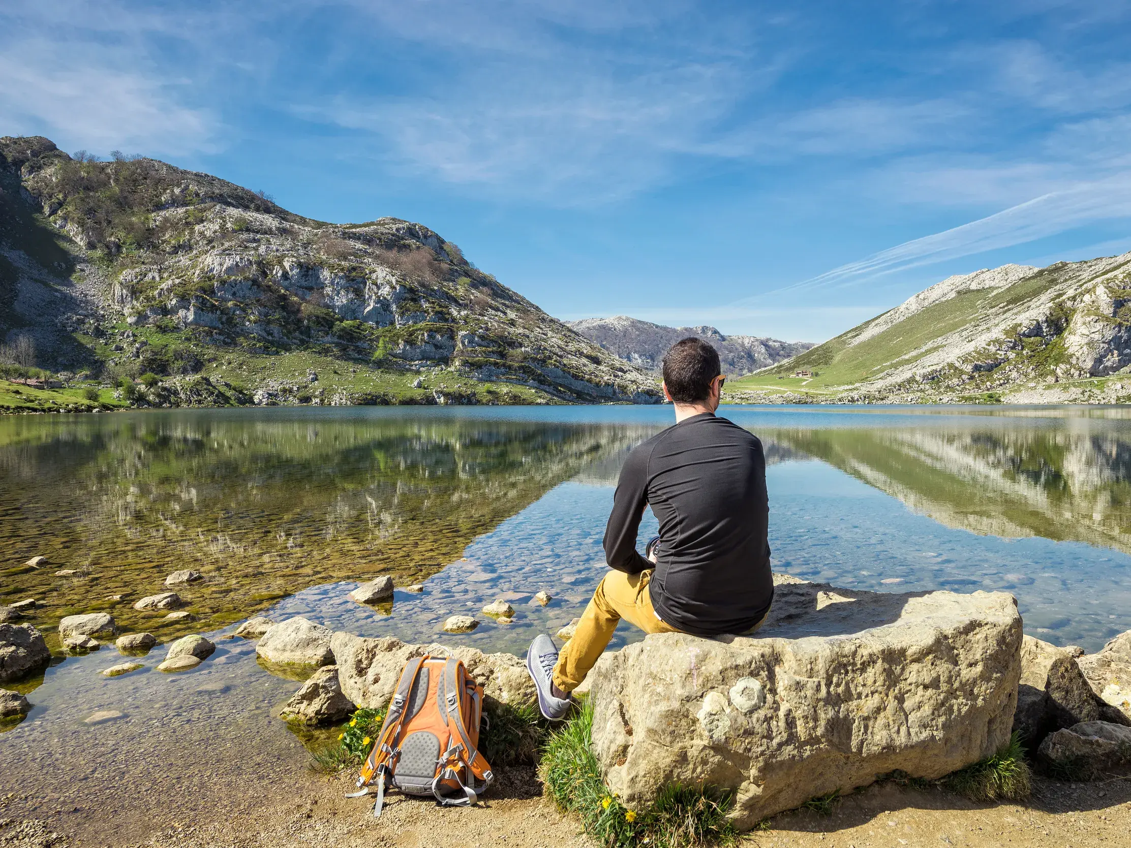 Ein Wanderer genießt bei einer Wanderreise auf einem Felsen sitzend den Ausblick auf einen klaren Bergsee in den Pyrenäen.