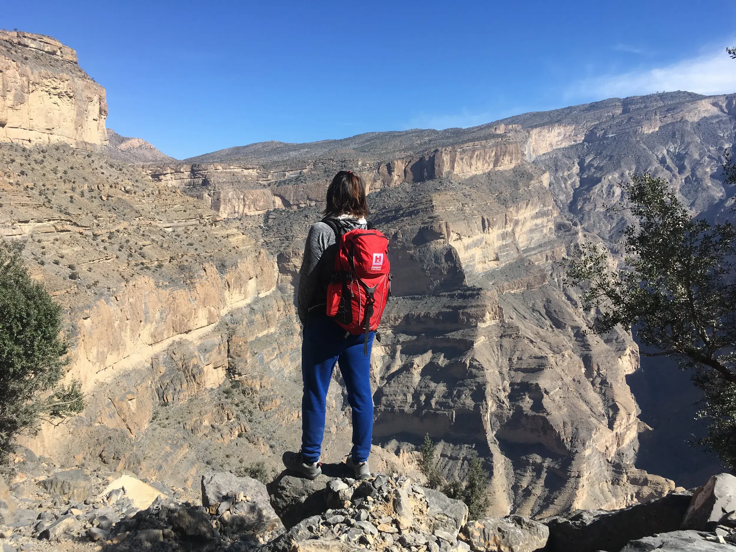 Eine Wanderin mit rotem Rucksack steht auf dem Balcony Walk am Jebel Shams und blickt in die tiefe Schlucht des Wadi Ghul im Oman.