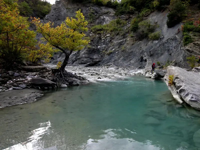 Zwei Trekker wandern am Ufer eines türkisfarbenen Wildflusses durch die unberührte Berglandschaft des Zagoria-Tals in Albanien.
