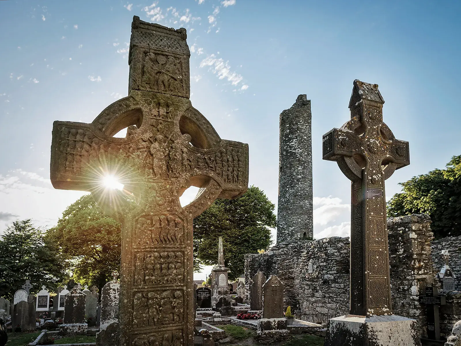 Die Sonne scheint durch ein steinernes Hochkreuz in der Klosterruine von Monasterboice in Irland, im Hintergrund ein Rundturm.