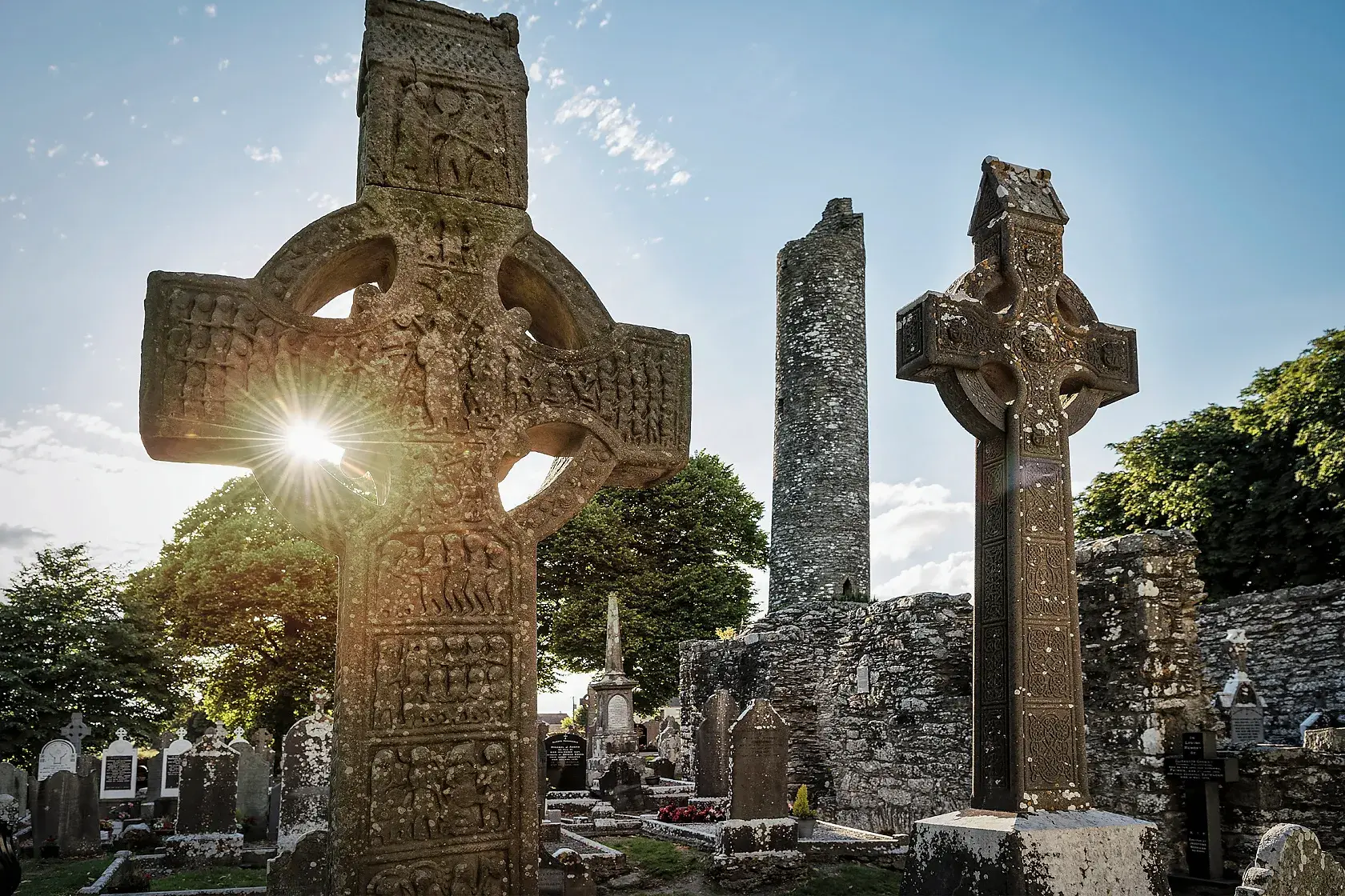 Die Sonne scheint durch ein steinernes Hochkreuz in der Klosterruine von Monasterboice in Irland, im Hintergrund ein Rundturm.