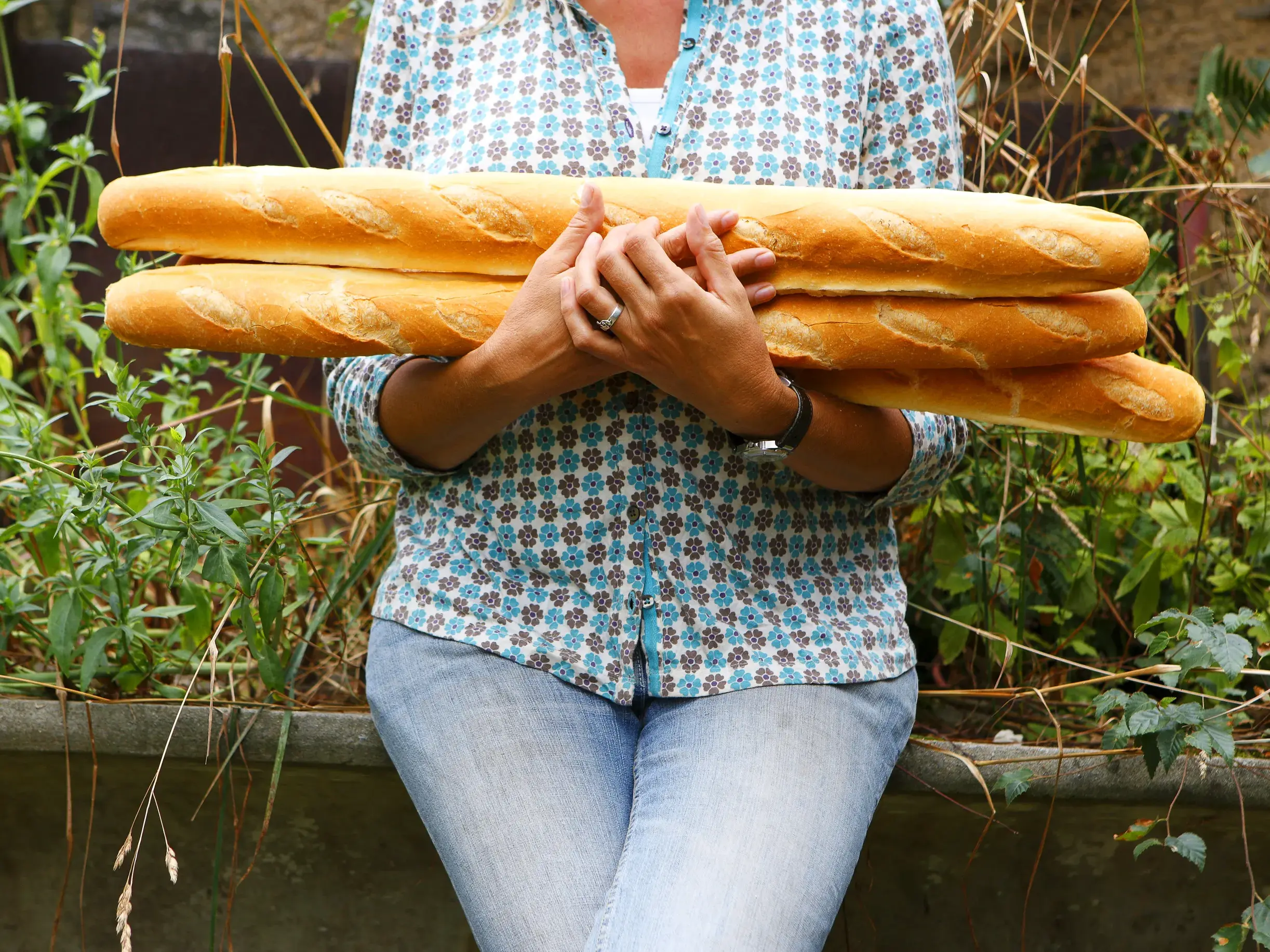 Eine Frau in Jeans und Bluse hält drei frische Baguettes in ihren Armen, ein typischer Genuss für ein Picknick im Périgord.