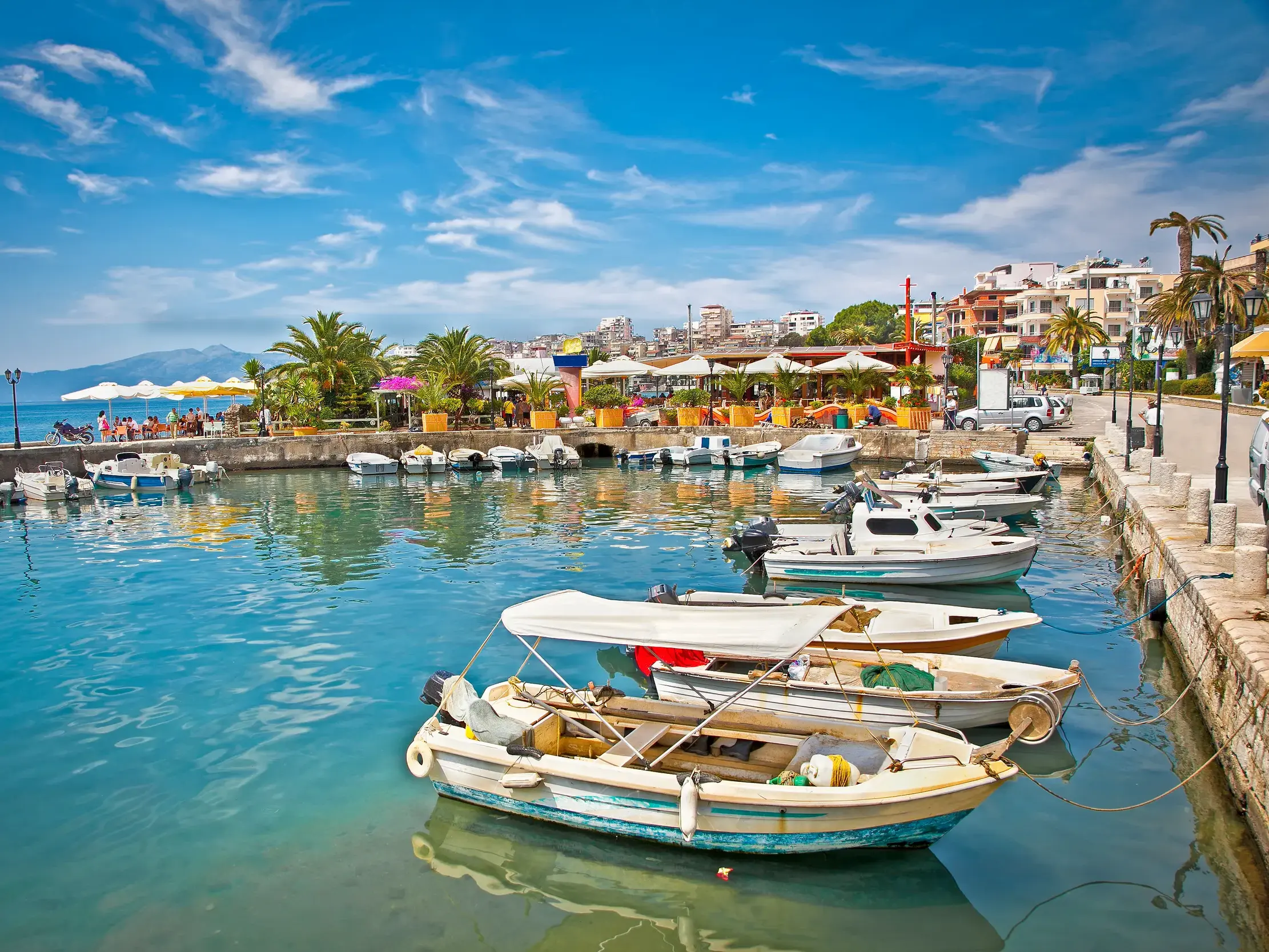Sonnige Uferpromenade im Hafen von Saranda mit kleinen Booten im türkisfarbenen Meer und Cafés am Ufer.