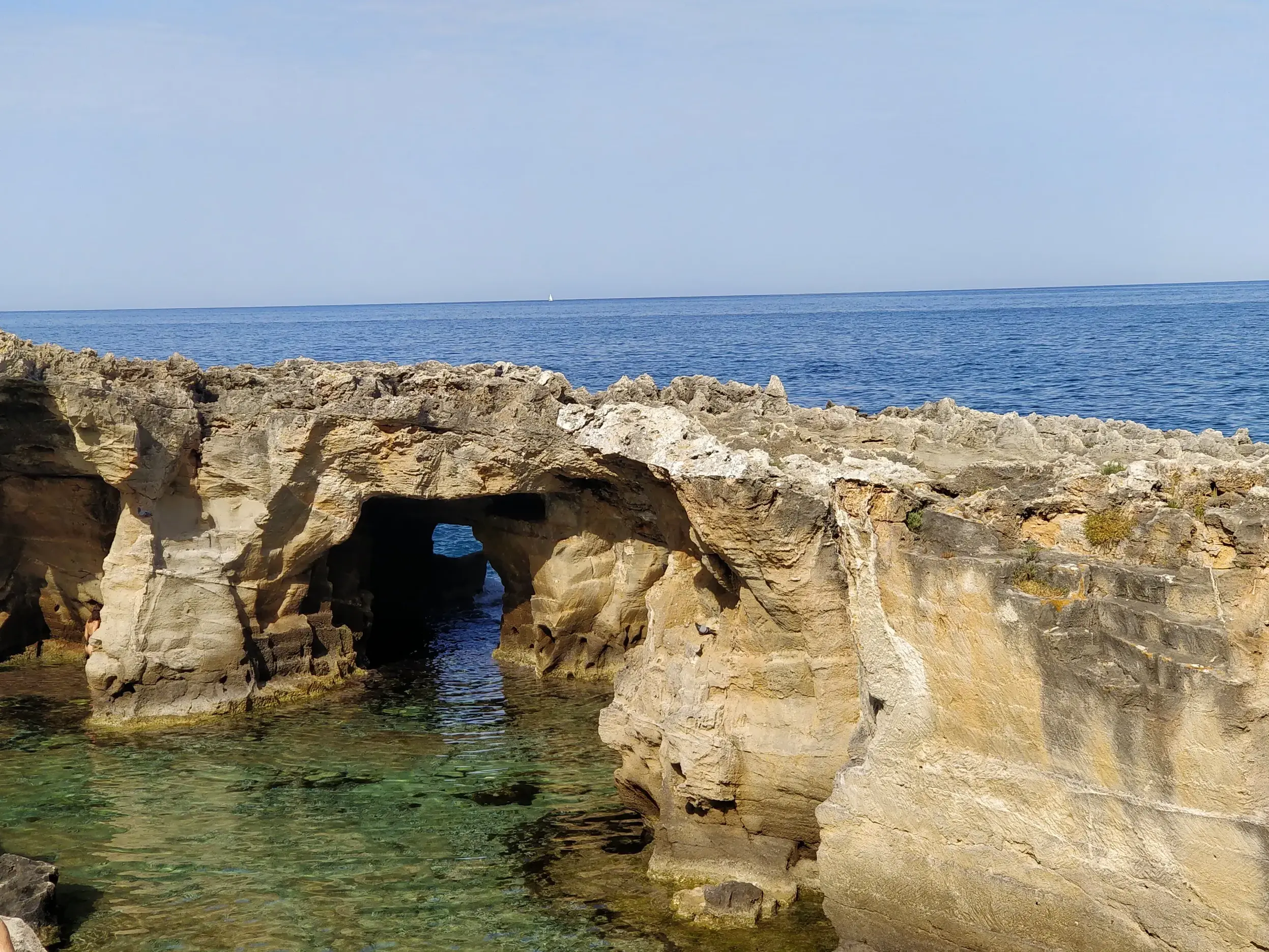 Zerklüftete Felsen mit Höhlen und einem Felsentor an einer Bucht mit türkisfarbenem Meer an der Küste Apuliens im Salento.
